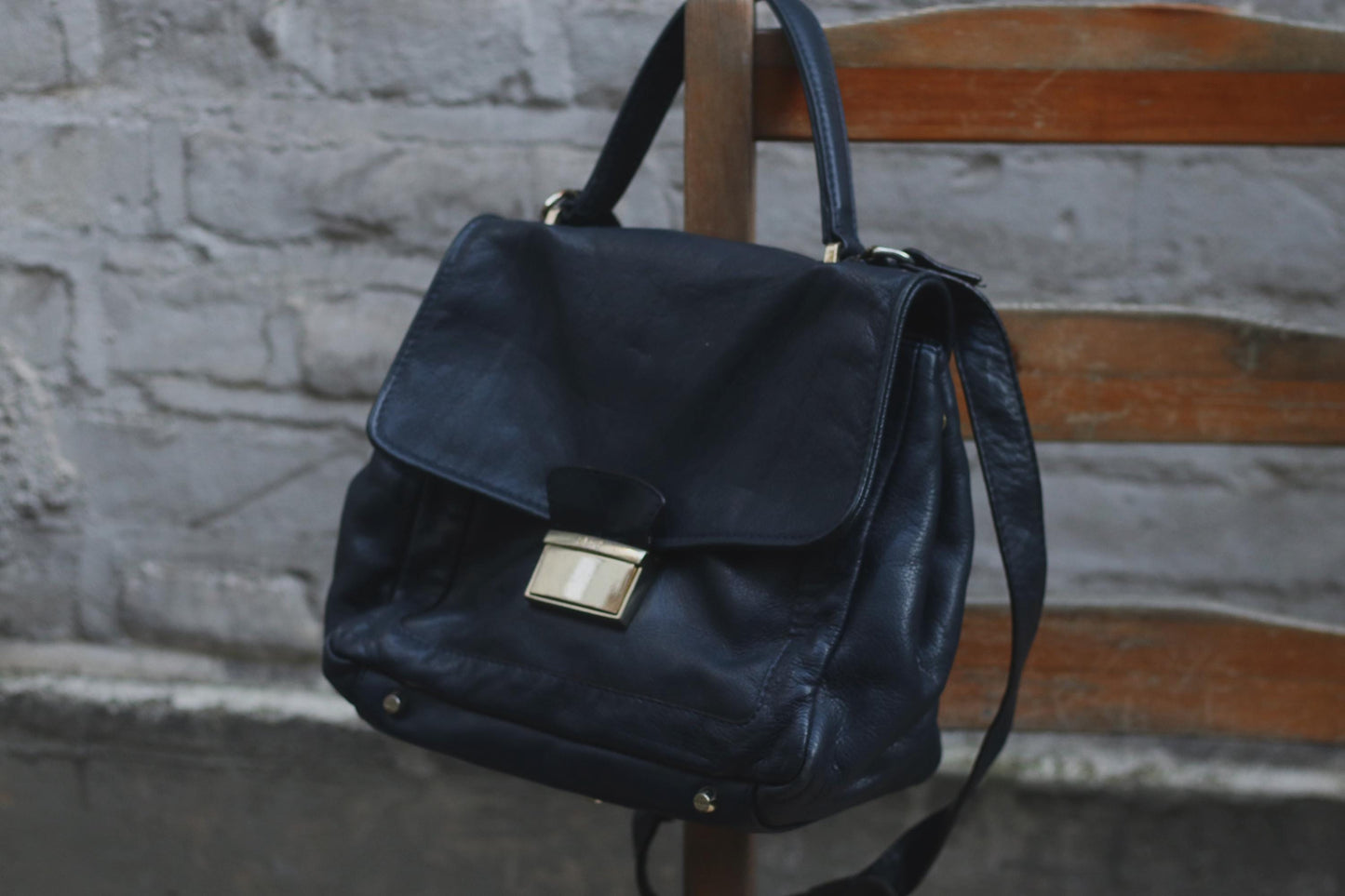 90s vintage black leather shoulder/crossbody bag with a top handle hanging from a wooden chair. The supple leather, classic flap shape, top handle, and crossbody strap make it a versatile accessory. A grey textured wall provides a simple backdrop.