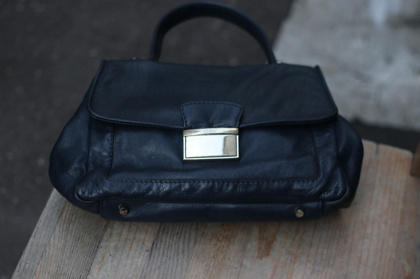 90s Black leather top-handle bag by Abro with silver-toned hardware on a wooden surface. Classic design featuring a flap closure and visible stitching. Soft, worn texture suggests vintage quality. Focus on the details and craftsmanship.