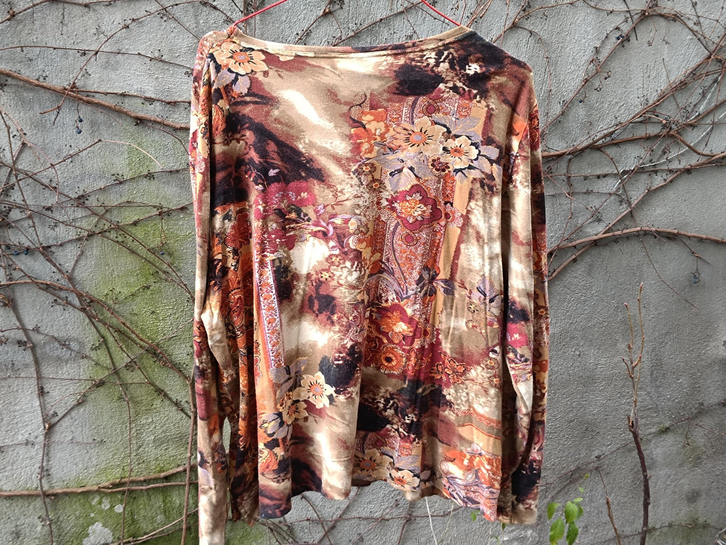 Vintage floral blouse in taupe, rust, and peach tones. Abstract floral print, boho style, round-neck, and lightweight. it's on a hanger against a textured grey and green background.