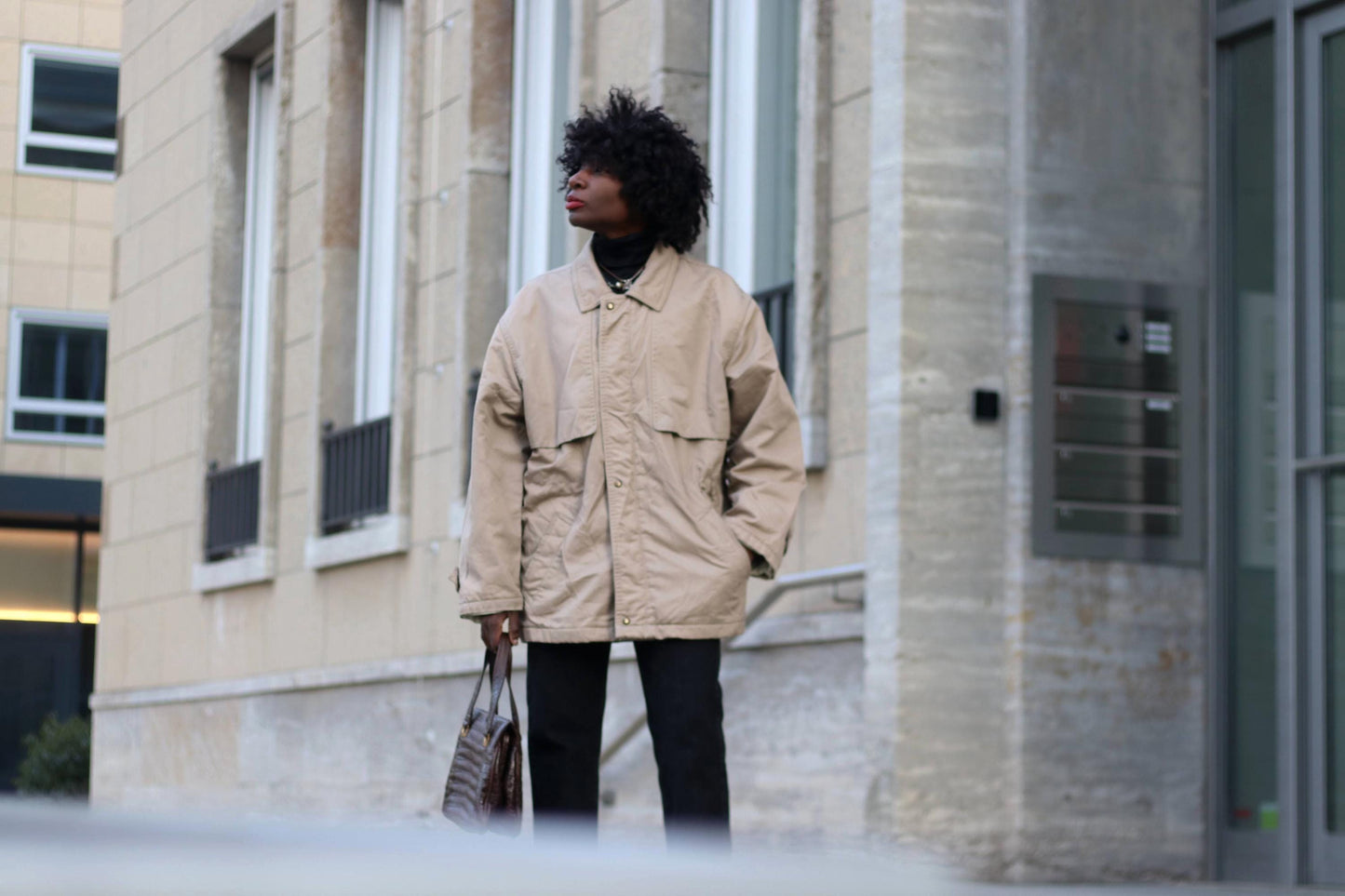 A stylish woman wearing a vintage oversized beige utility jacket with a relaxed fit, structured shoulders, and large pockets, standing confidently outside a modern concrete and glass building.