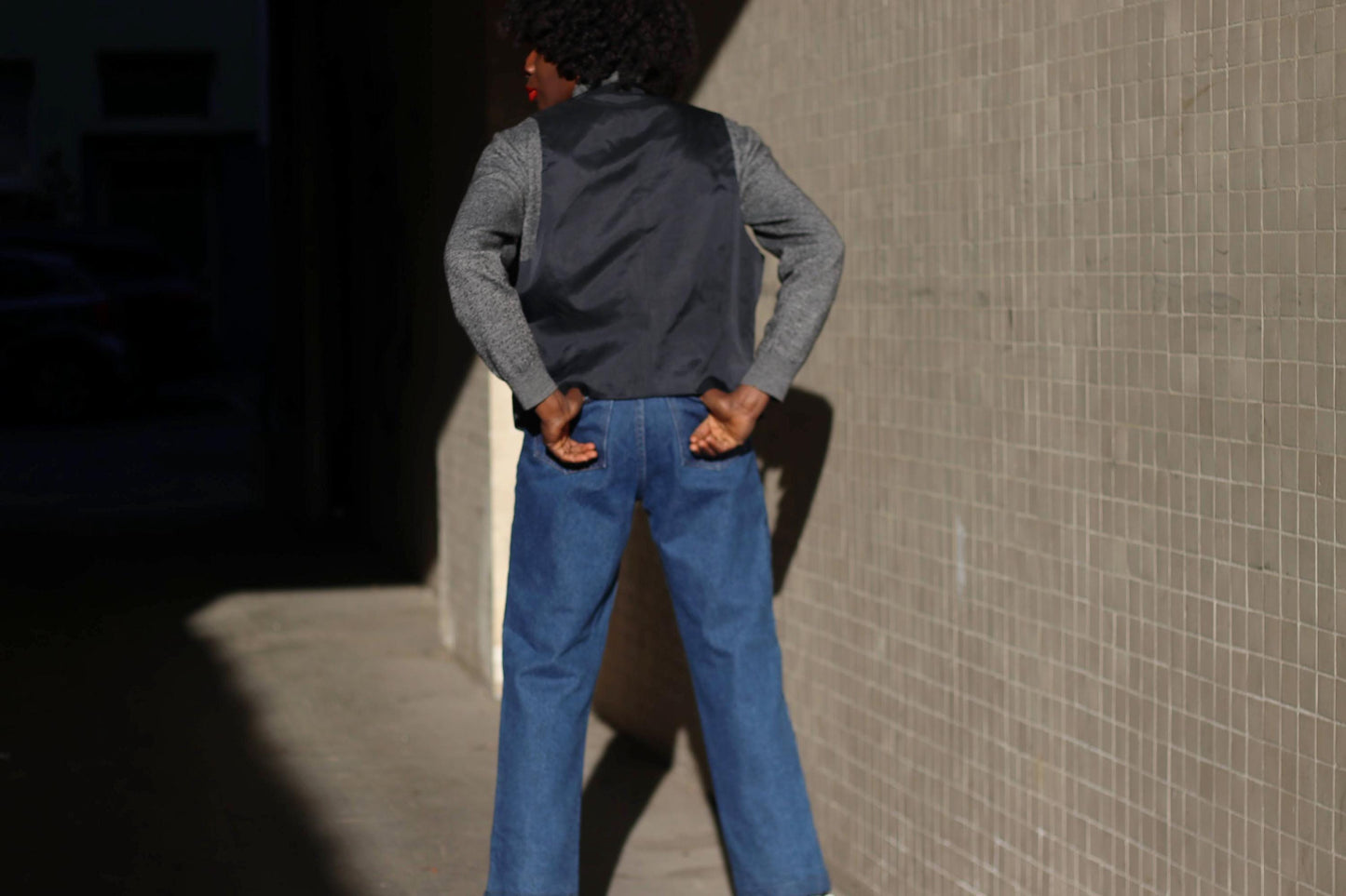 A model showcasing the back of a vintage 90s tartan plaid vest in navy and green over a gray turtleneck, styled with blue jeans, standing against a textured urban wall