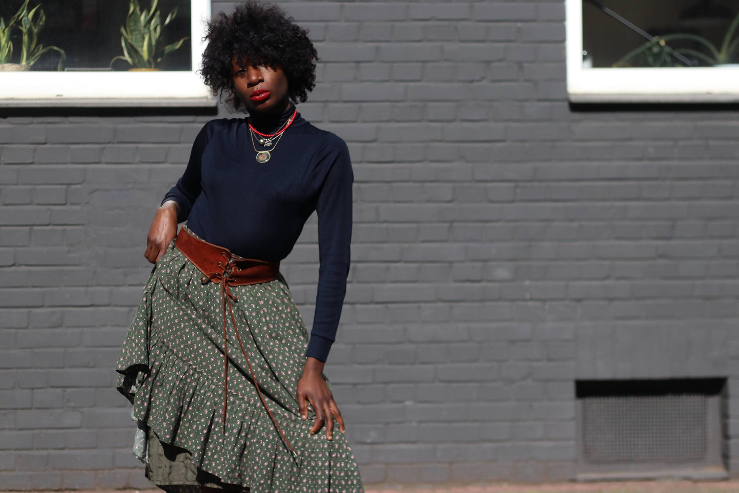 A female model wearing a vintage 70s green midi skirt with an intricate eyedrop print and ruffle hem, styled with a boho belt, standing against a gray brick wall.