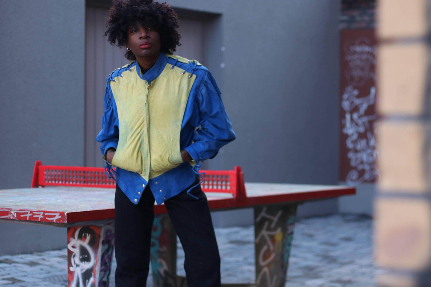 A woman with curly black hair and bold red lipstick wearing a vintage 80s suede jacket with bold blue and yellow colorblocking, standing in front of a graffiti-covered ping pong table photographed with her hands in her pocket.