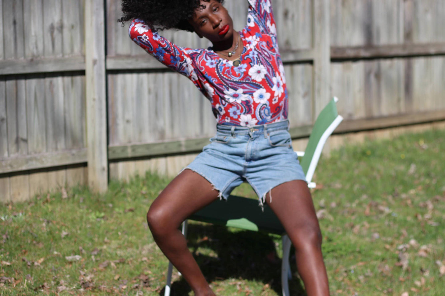 A female model with curly black hair sitting on a white and green lawn chair on a sunny lawn, showcasing a red, white, and blue patterned vintage Talbot's blouse made of 100% merino wool. A wooden fence and green foliage form the backdrop. The model has her hands in her hair