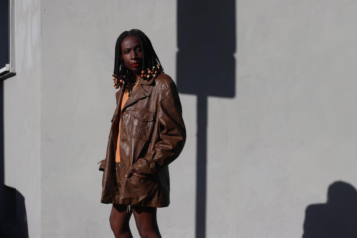 Female model photographed in a 90s brown Leather Jacket infront of a white wall with a shadow of a street sign. The unisex jacket is styled with an orange v-neck sweater and blue denim shorts.