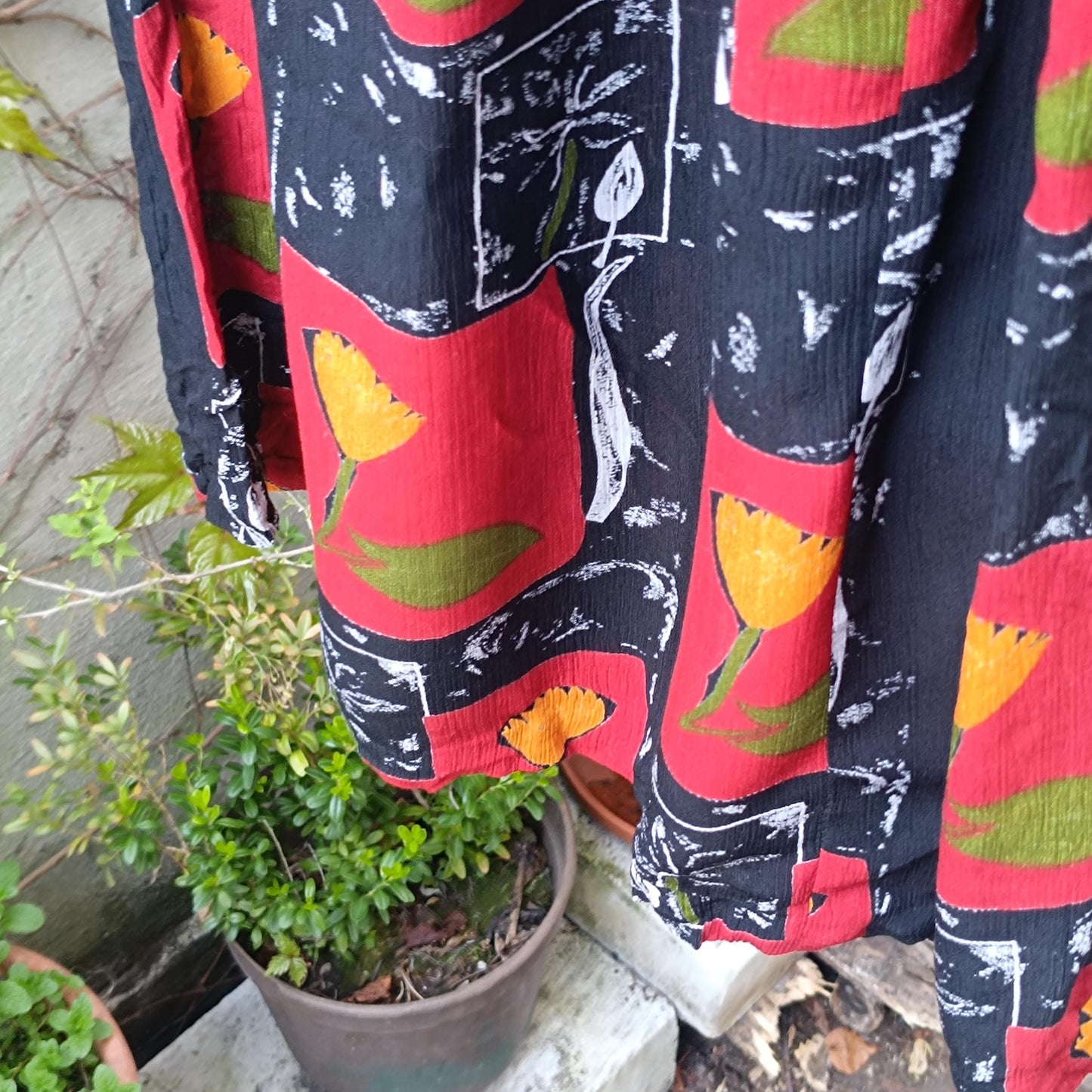 A still life image shot outdoors in a garden The picture is a Close-up image of a vintage 90 maxi skirt’s floral pattern showing red blocks with yellow flowers and leafy green accents, emphasizing the retro boho print and summer vibe.