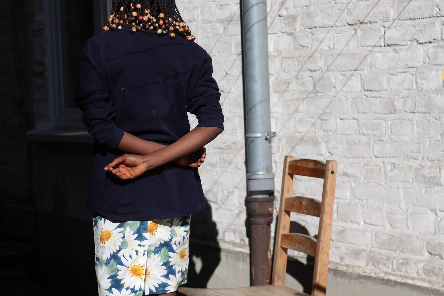 A back view of a female model wearing a dark long-sleeved shirt and a white knee-length skirt with a blue and yellow daisy pattern, standing next to a wooden ladder