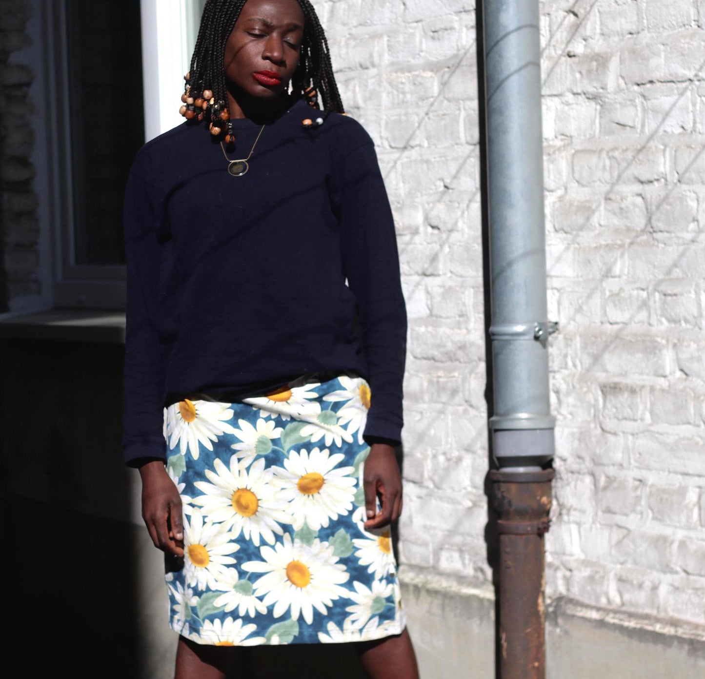 Female model in a 90s vintage daisy print knee-length skirt in white and blue paired with a dark blue sweater is photographed in front of white wall with lined shadows