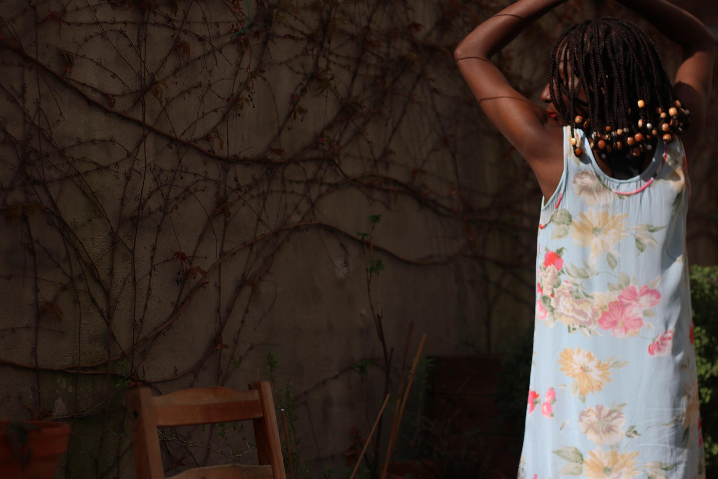 An ethereal grunge dress from the 90s with a sky blue floral print and layered hem is shown on a female model outdoors against a wall with climbing vines and a rustic wooden chair. The model's back is to the camera