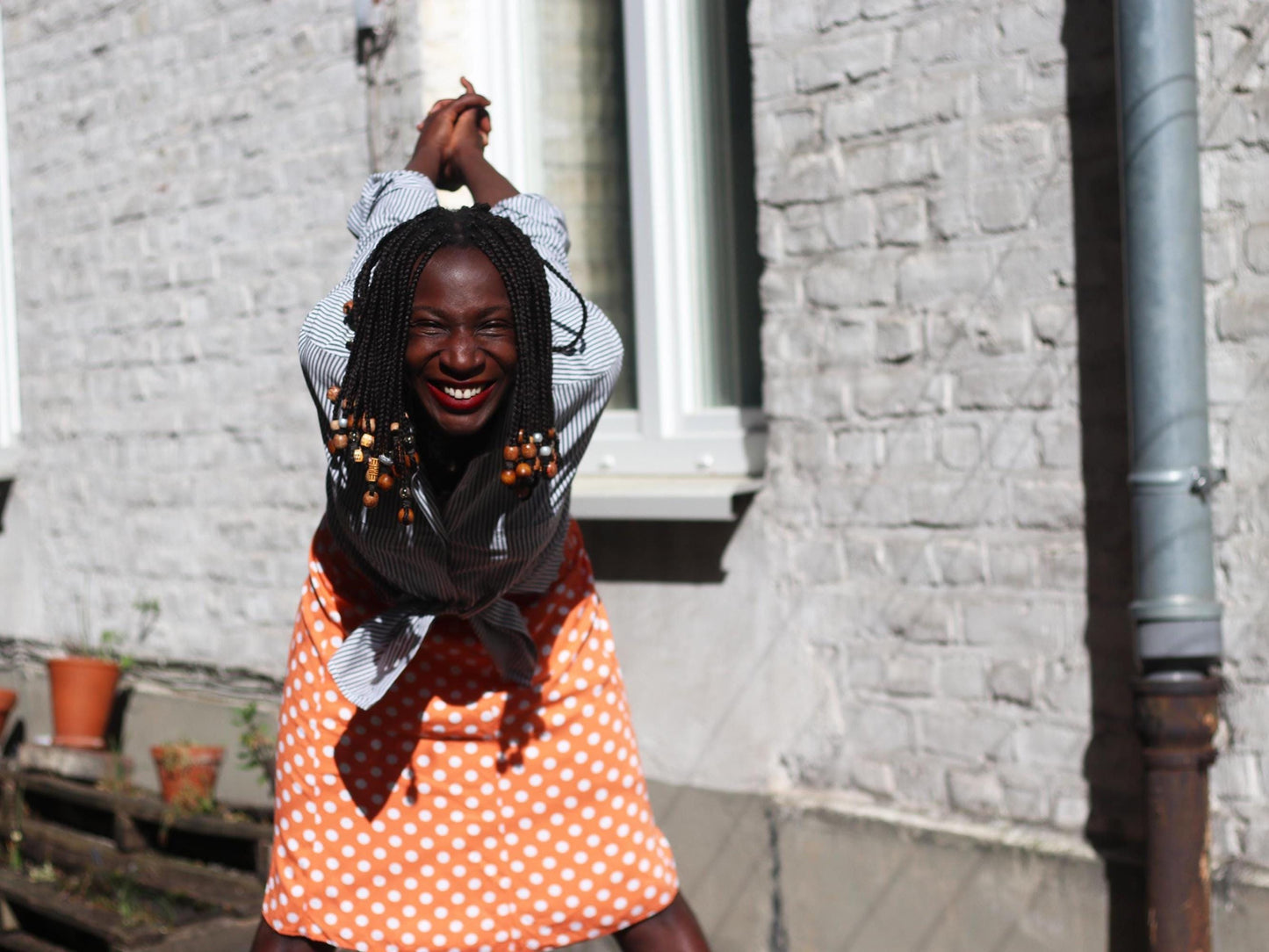 A smiling female model in a Chic French Flair 90s Polka polka-dotted skirt paired with striped shirt. The vintage orange skirt exudes a flirty Parisian Summer vibe and style. The model has her hands are behind her back in a playful manner