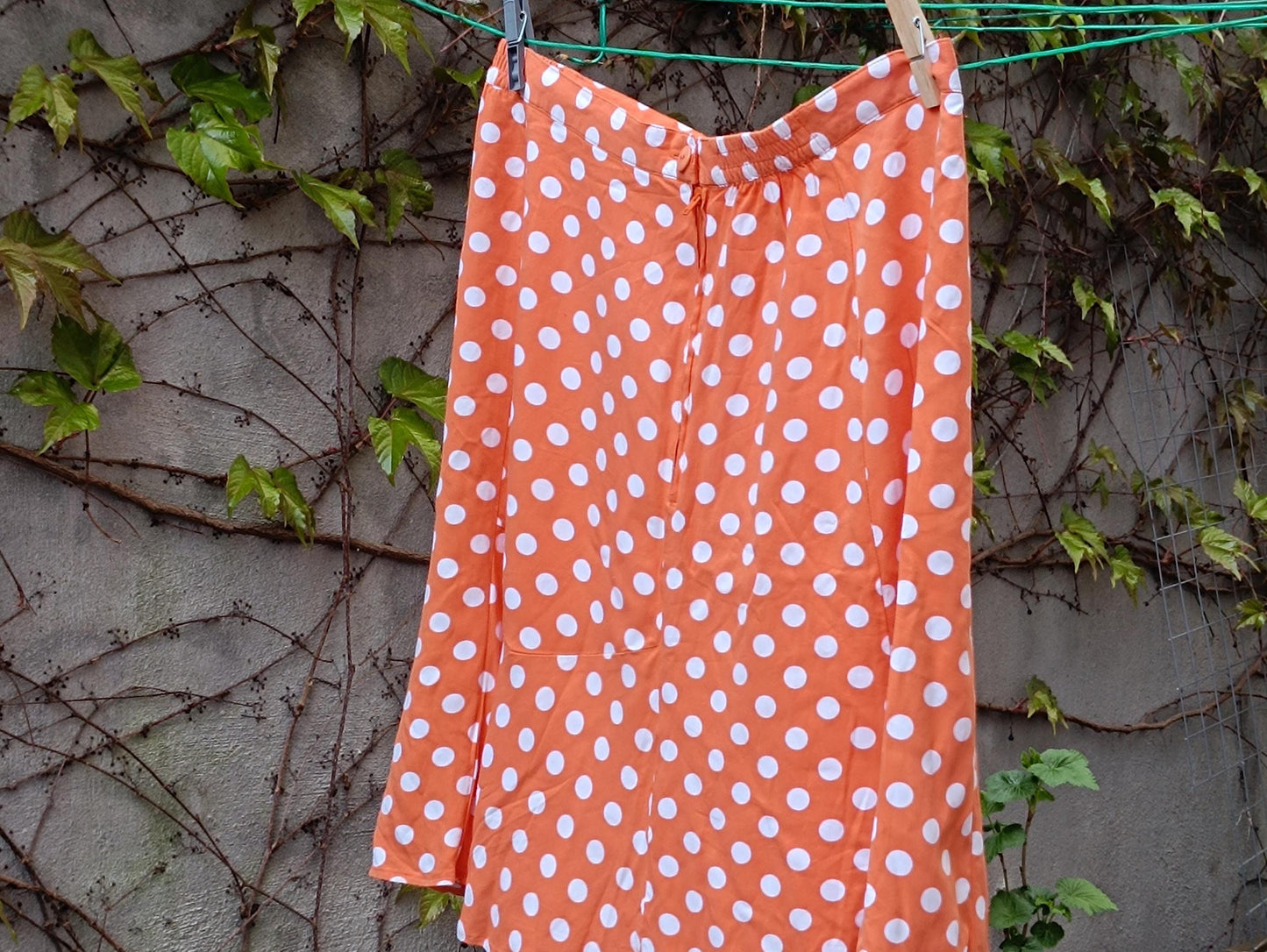 A Chic French Flair 90s Polka Dotted Skirt in Orange is captured on a clothing line outdoors in a garden against a dreamy wall with leavy vines. The vintage Flirty Mini Skirt excuses a Parisian Summer vibe