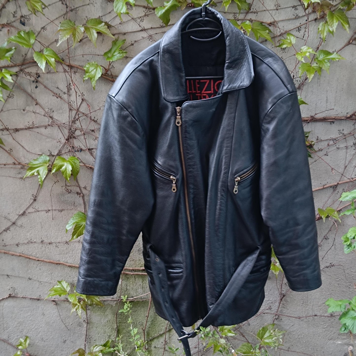 Vintage 90s Collezione Del Tempo black leather jacket photographed outdoors hanging against a grey wall with green plants