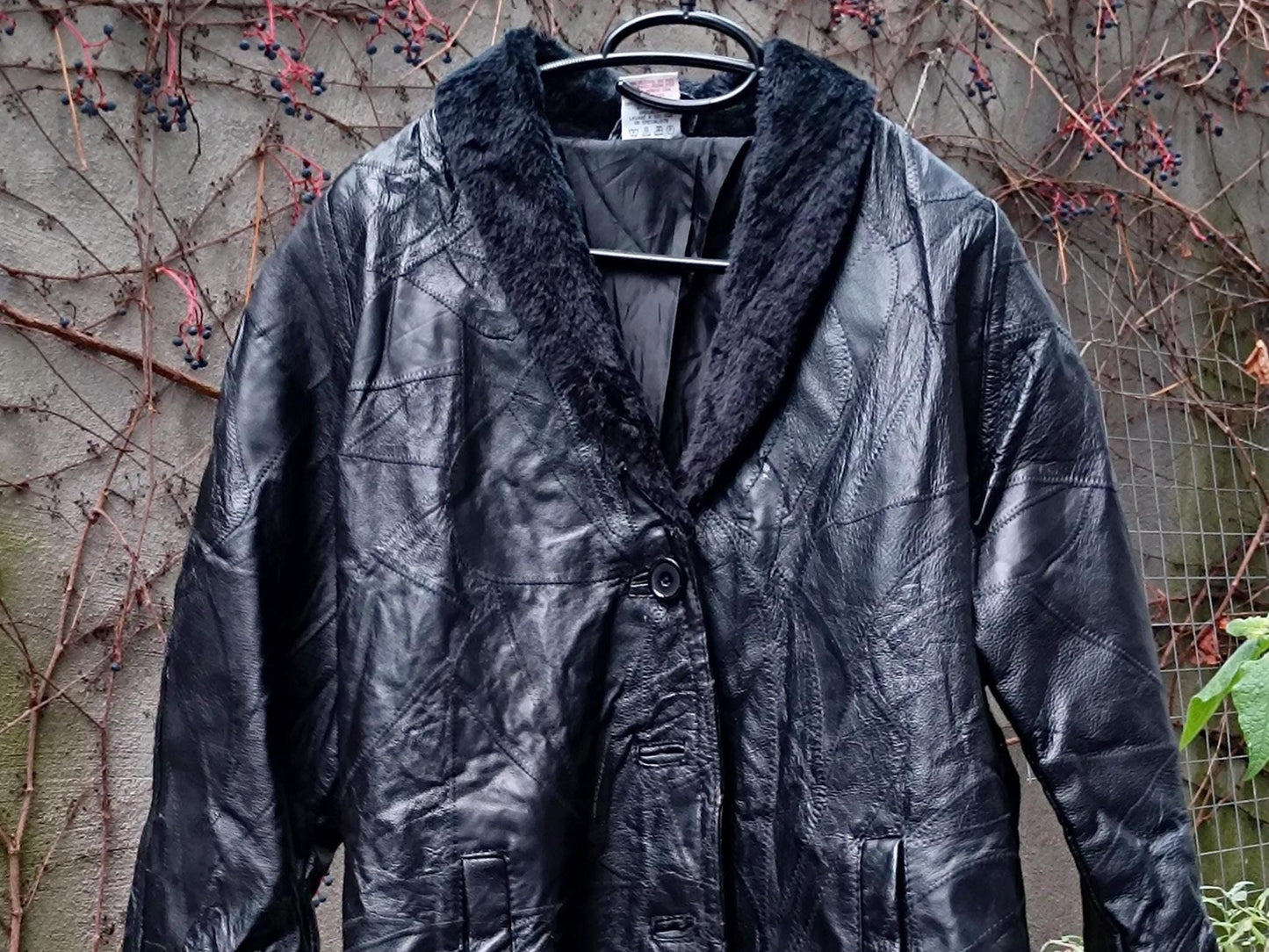 A vintage black leather jacket with a faux fur trimmed coat by the brand Anne De Lancay Women's Coat photographed hanging outdoors on a clothing line against a grey wall with browning twines and leaves