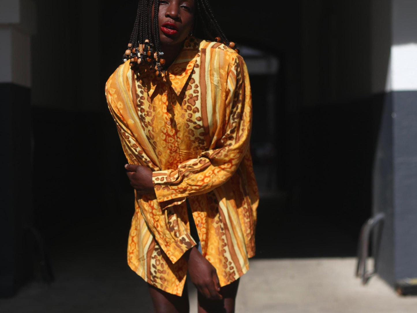 Outdoor shot of a person with beaded braids wearing a yellow and brown patterned long blouse and jeans, photographed standing outdoors. The model looks directly into the camera with one hand touching the elbow other other arm. The focus is on the oversized vintage 90s shirt.