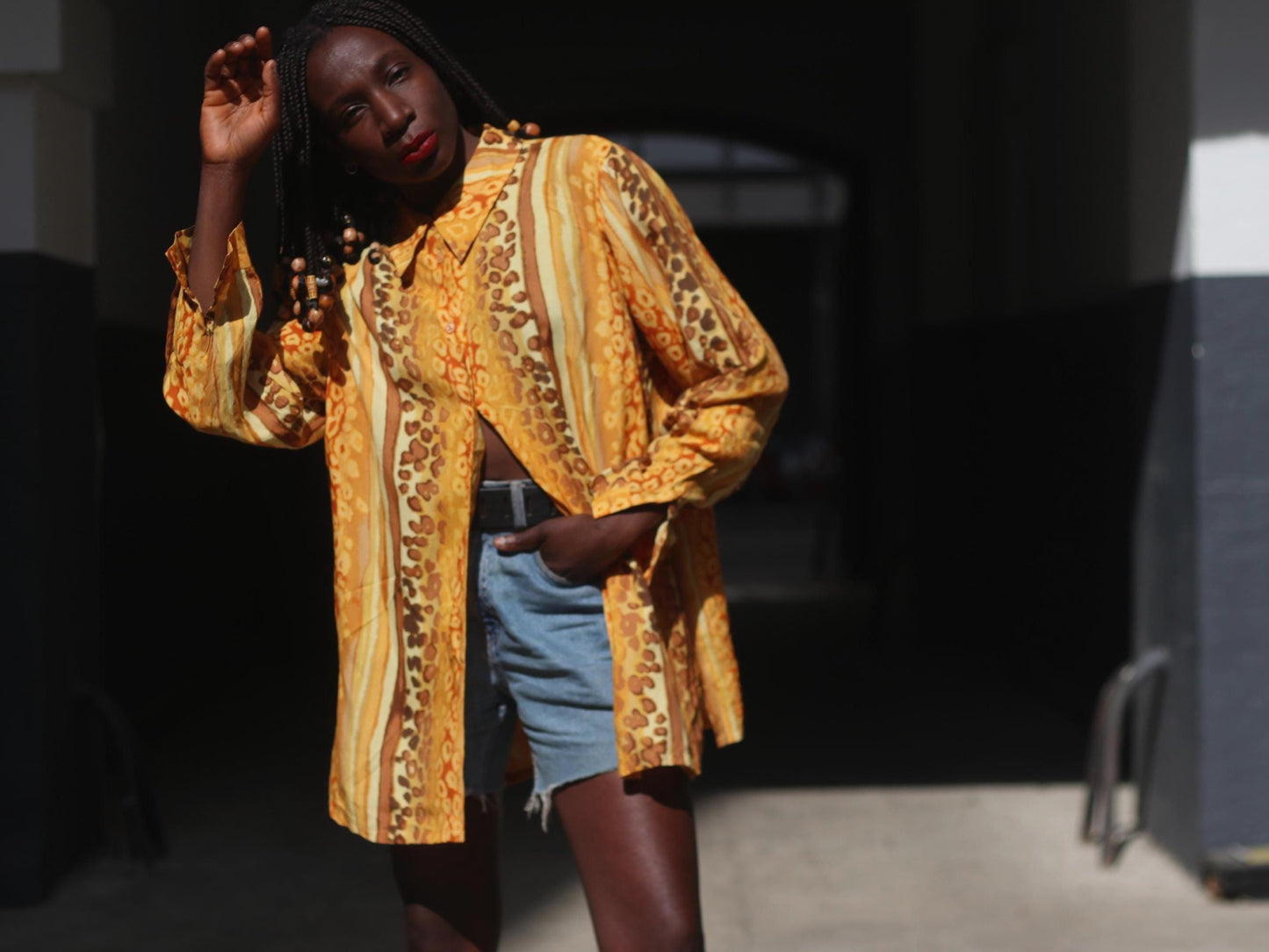 A woman with braided hair with beads modeling a vibrant yellow vintage blouse. The 90s blouse features a bold animal print with swirling patterns in shades of yellow, gold, brown, and cream. It has long sleeves, a button-down front, and a loose, oversized fit. She pairs it with light blue denim shorts. She lok=oks directly into the camera with one hand raised.