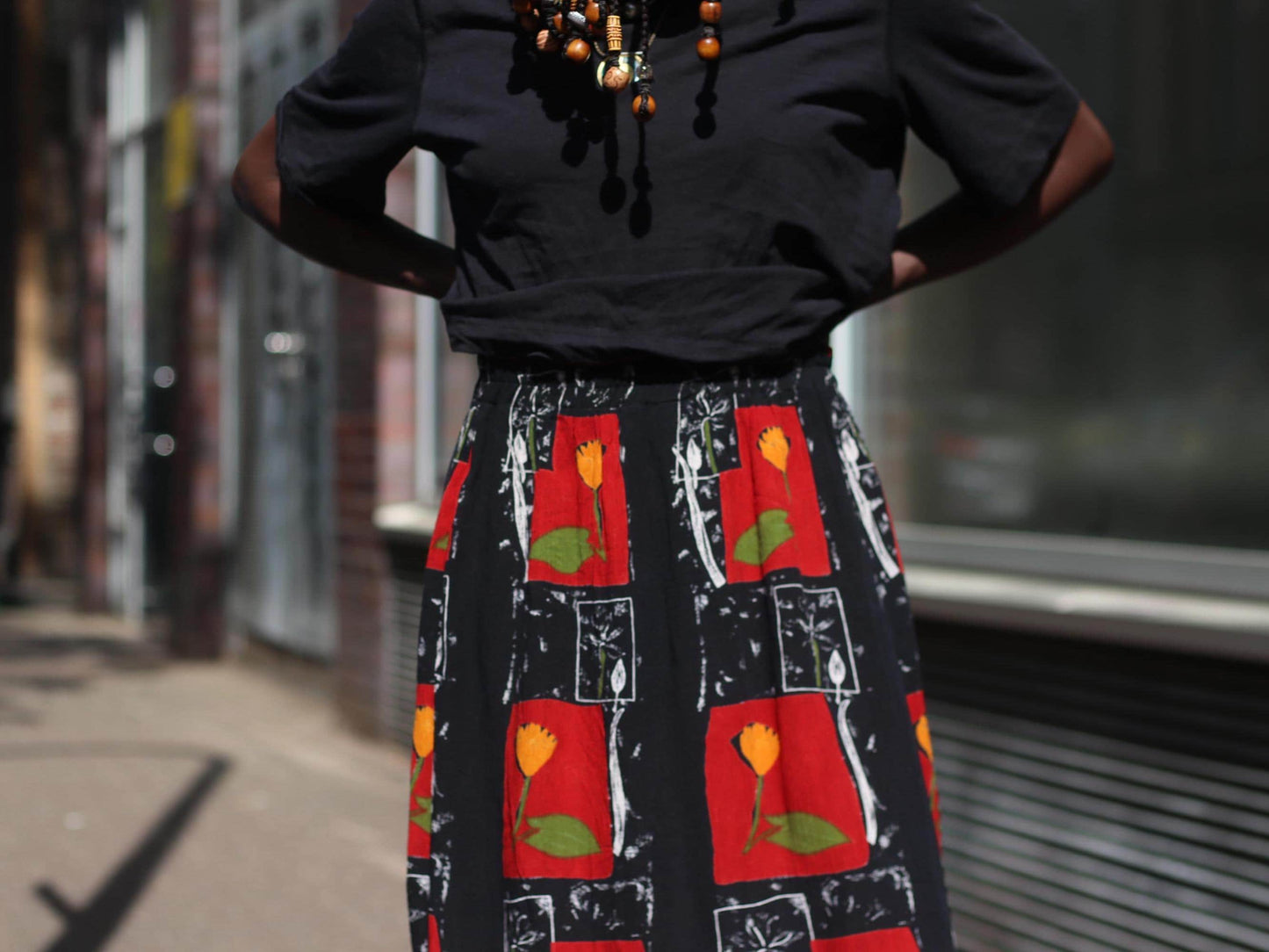 A close-up view of a vintage skirt’s floral pattern showing red blocks with yellow flowers and leafy green accents, emphasizing the retro boho print and summer vibe. The 90s skirt is showcased on a female model whose face is unseen. Her braided hair is seen