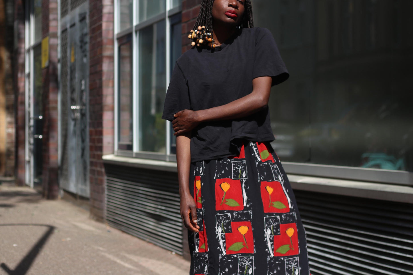 A female model in a calm pose with on arm touching the elbow of the other, wearing a striking 90s red floral vintage maxi skirt, surrounded by urban architecture and soft afternoon sunlight.