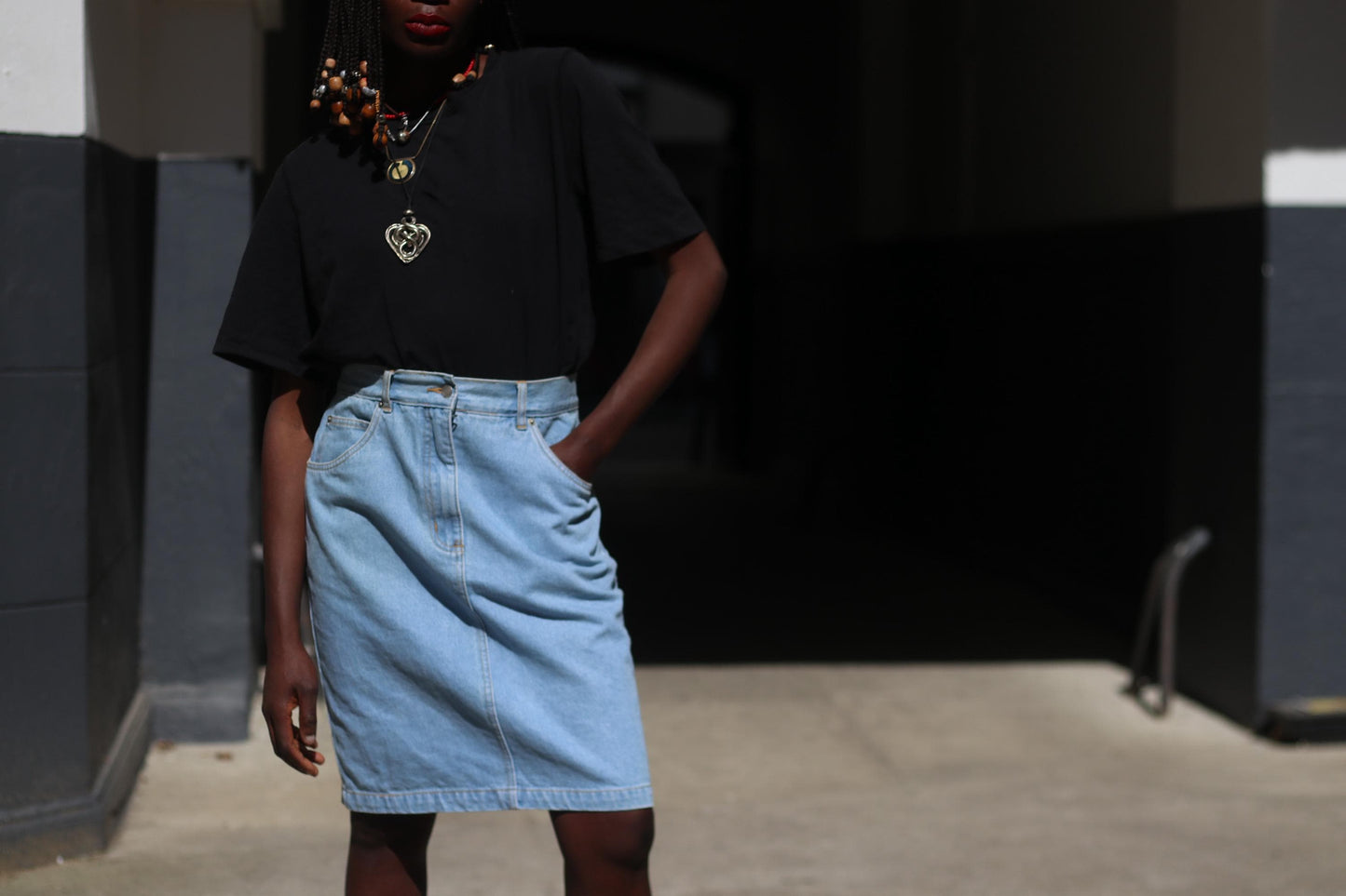 A front view of a vintage 90s blue denim skirt showcased on a female model with beaded braids shot outdoors in a sunlight alley. Model wears the classic Knee-Length Jeans Skirt with a black and has one hand in her pocket