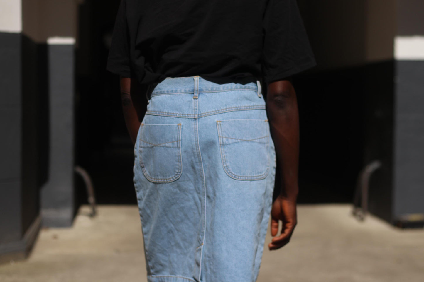 Back view of a vintage 90s blue denim skirt showcased on a female model shot outdoors in a sunlight alley. Model wears the classic knee-Length Jeans Skirt with a black t shirt. The focus is on the back pockets of the denim skirt and the stitchings