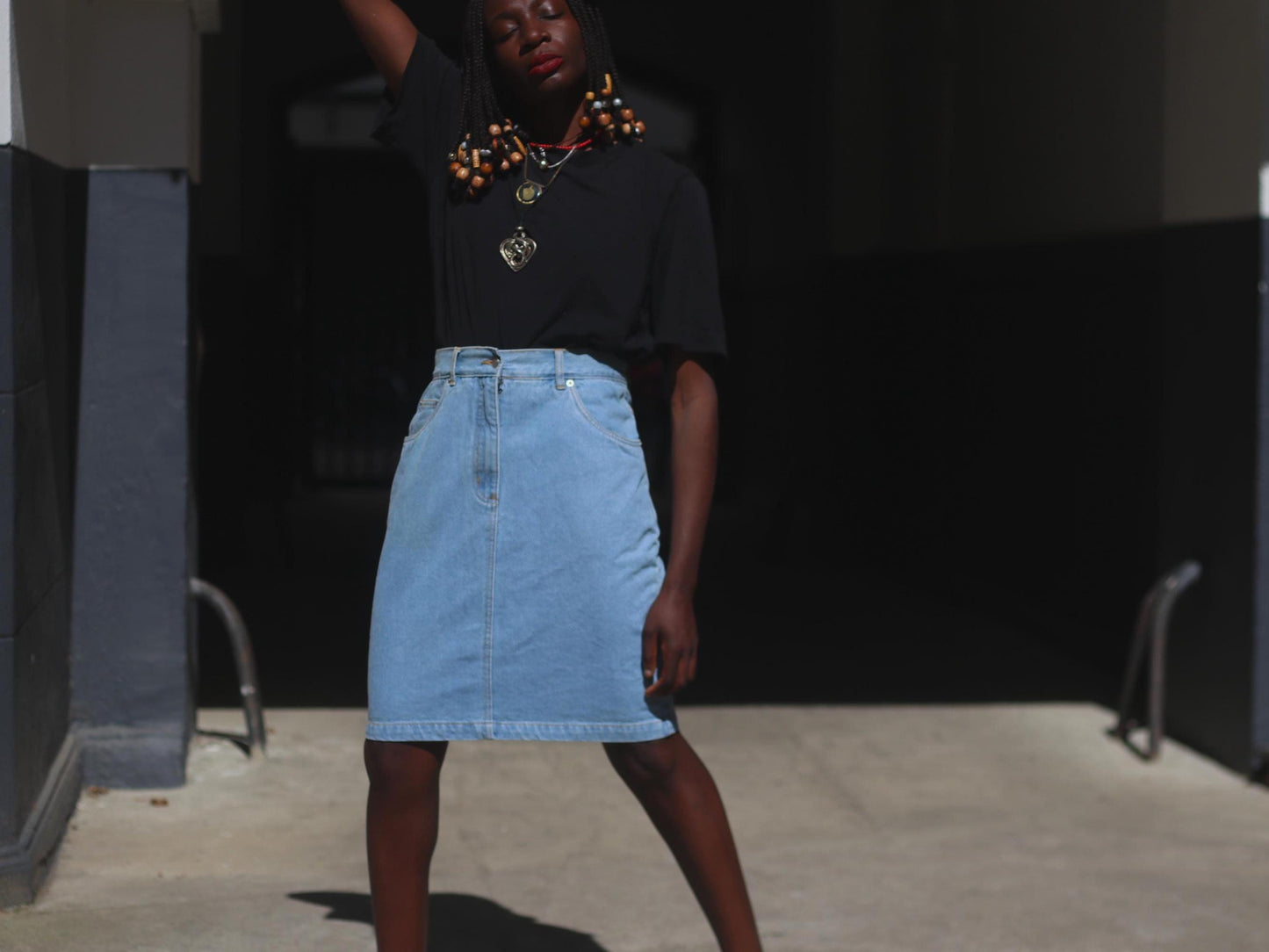 Vintage 90s blue denim skirt showcased on a female model with beaded braids shot outdoors in a sunlight alley. Model wears the classic knee-length Jeans Skirt with a black t shirt.