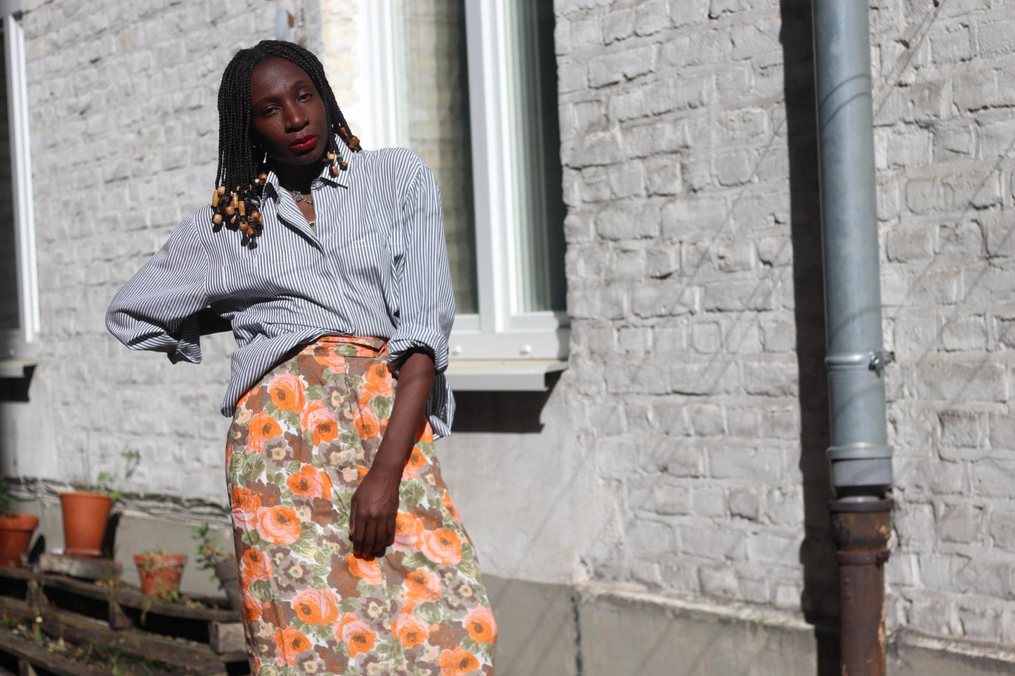 Female model wearing an 80s Floral Skirt in orange and brown styled with a pinstriped button-up in photographed outdoors against a sunlight white bricked wall