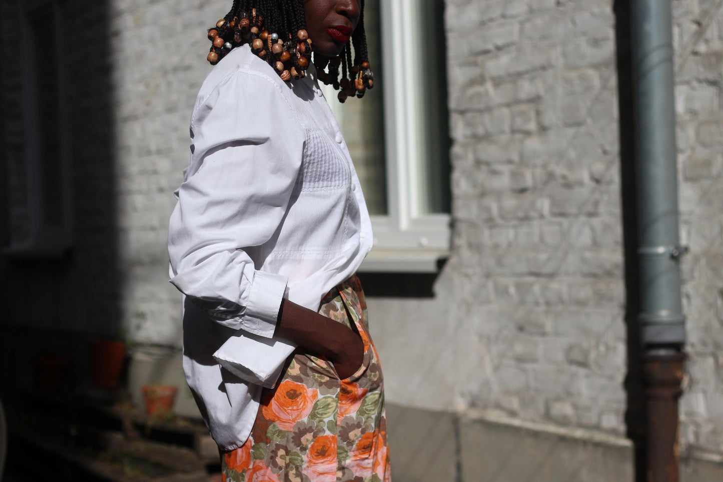 Detailed shot of a floral orange skirt styled with a pinstriped button-up in photographed outdoors against a sunlight white bricked wall. Model has one had in the pocket to show the practicality of the summer linen-blend skirt.