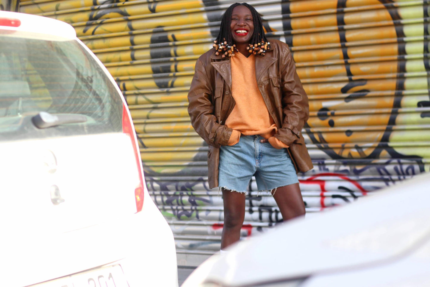 Female model photographed smiling in a 90s brown Leather Jacket infront of a graffiti covered wall. The unisex jacket is styled with an orange v-neck sweater and blue denim shorts. The jacket is unisex and minimalist in style. The vibe of picture is 90s cool street style