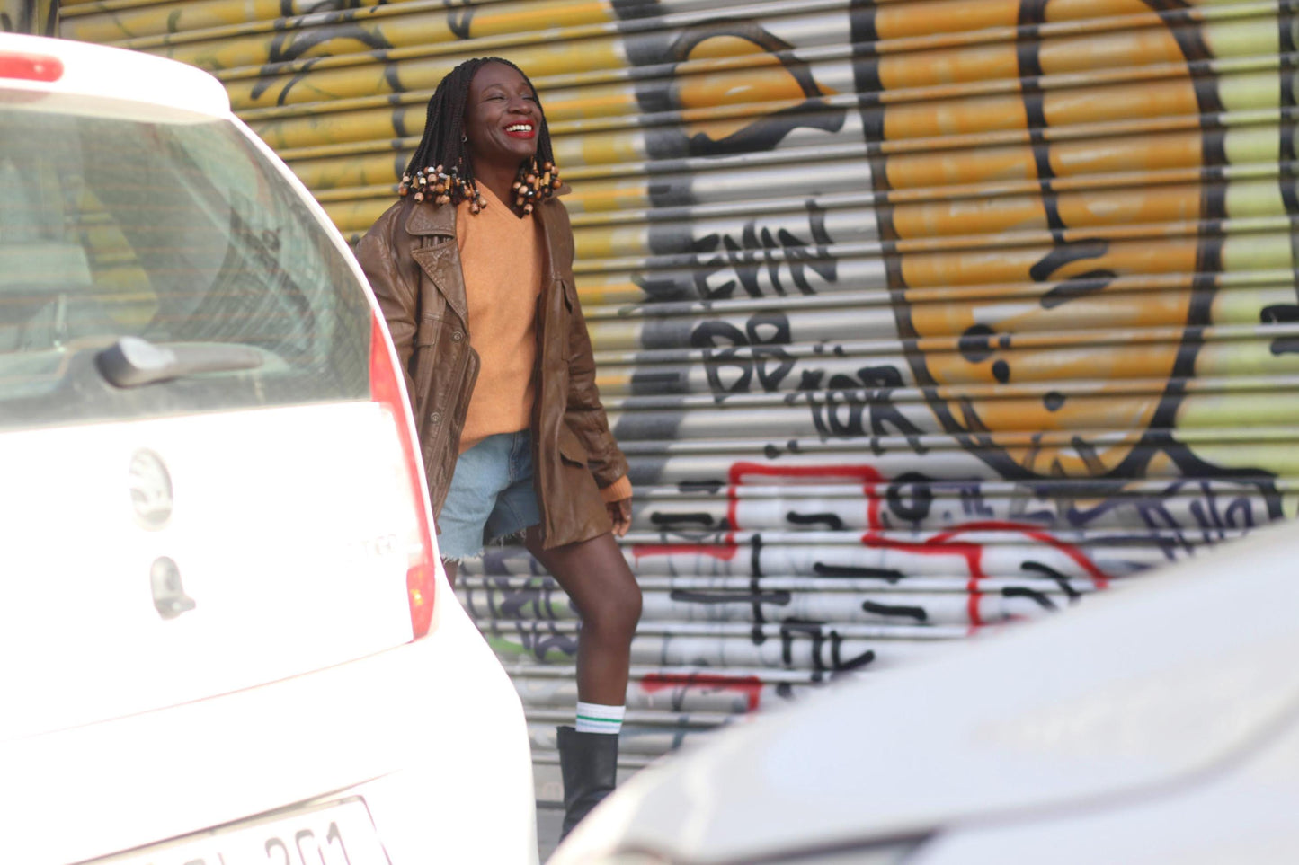 Side view of a female model photographed smiling in a 90s brown Leather Jacket infront of a graffiti covered wall. The model has braids with beads. The unisex jacket she is wearing is styled with an orange v-neck sweater, blue denim shorts and black boots with socks peaking out. The feel of image is 90s cool street style