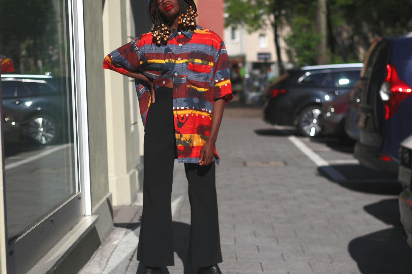 Sun lit photograph of a female model with beaded braids in a 90s vintage Montino tropical floral shirt with a retro bold island print, black pants and hat captured in an urban street setting with cars and buildings in the background. The image highlights the shirt's red and yellow hues, short sleeves, unisex fit, and bold summer tones.
