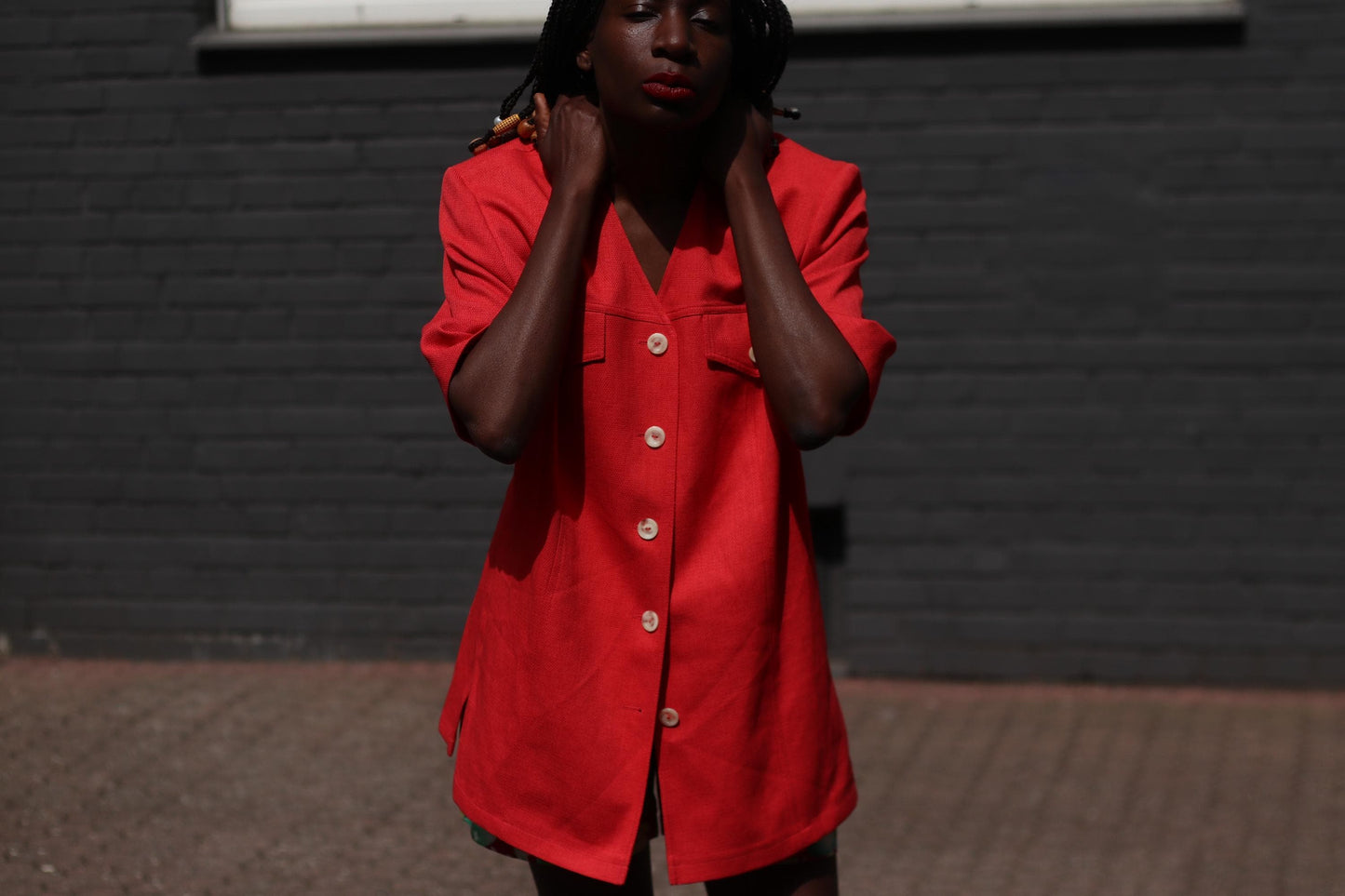 Female model in a vintage 90s Short-Sleeve Blazer by Mark Wald as a short dress photographed in front of a grey brick wall. The image showcases the standout features , from the v-Neck silhouette to the buttons and chest pockers