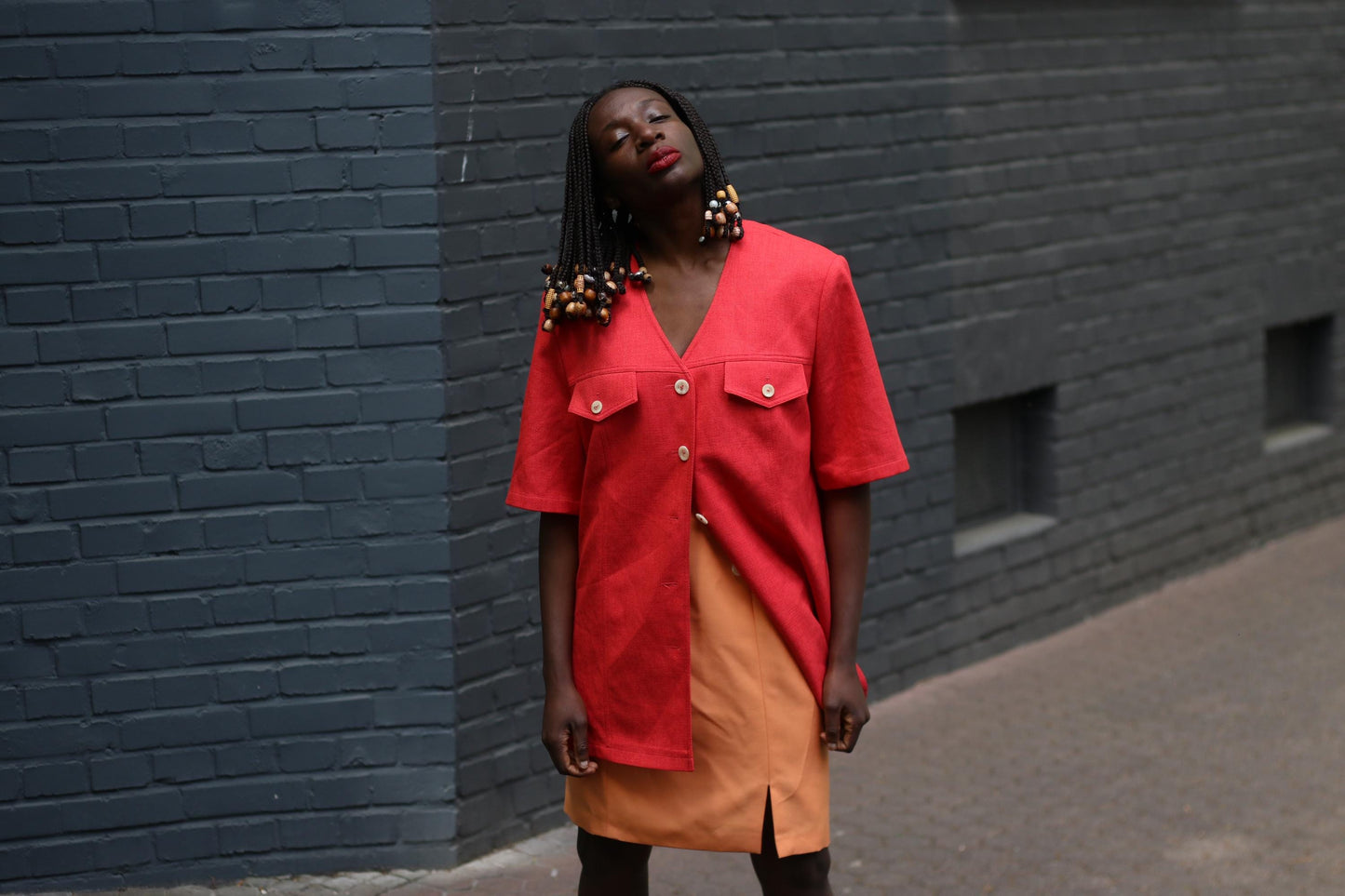 Female model with beaded braids wearing a vintage 90s Short-Sleeve Blazer by Mark Wald with a bold orange knee-length skirt photographed in front of a grey brick wall.