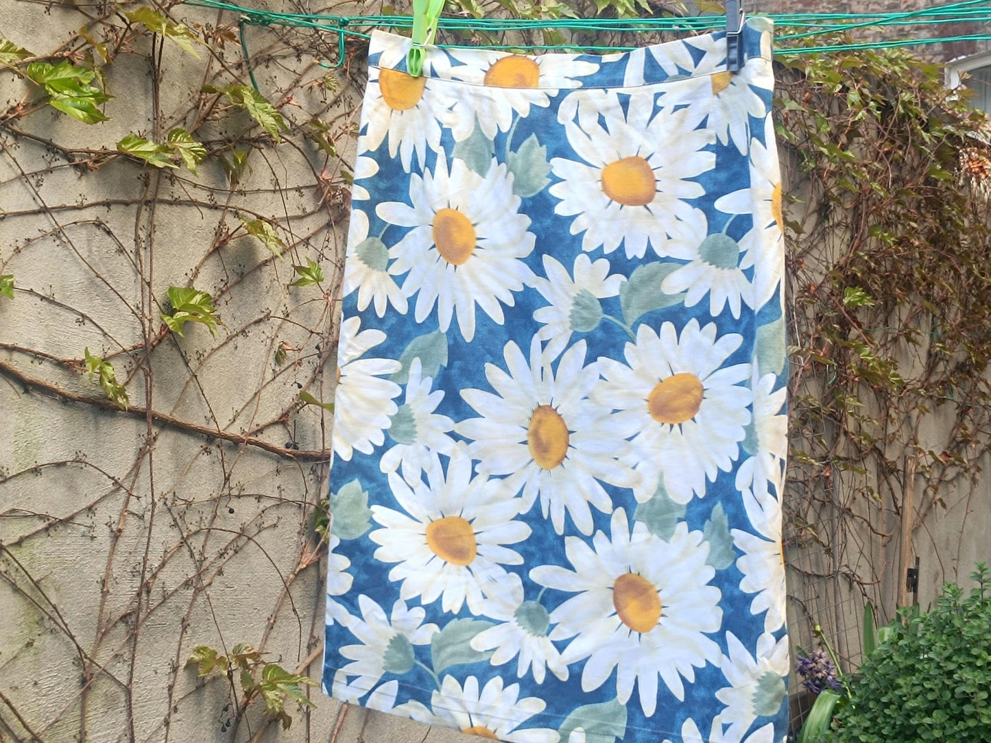 90s vintage blue and White Floral Print Skirt with a bold daisy print hanging on a clothing line in the garden against a walled background with browning ivy and twines