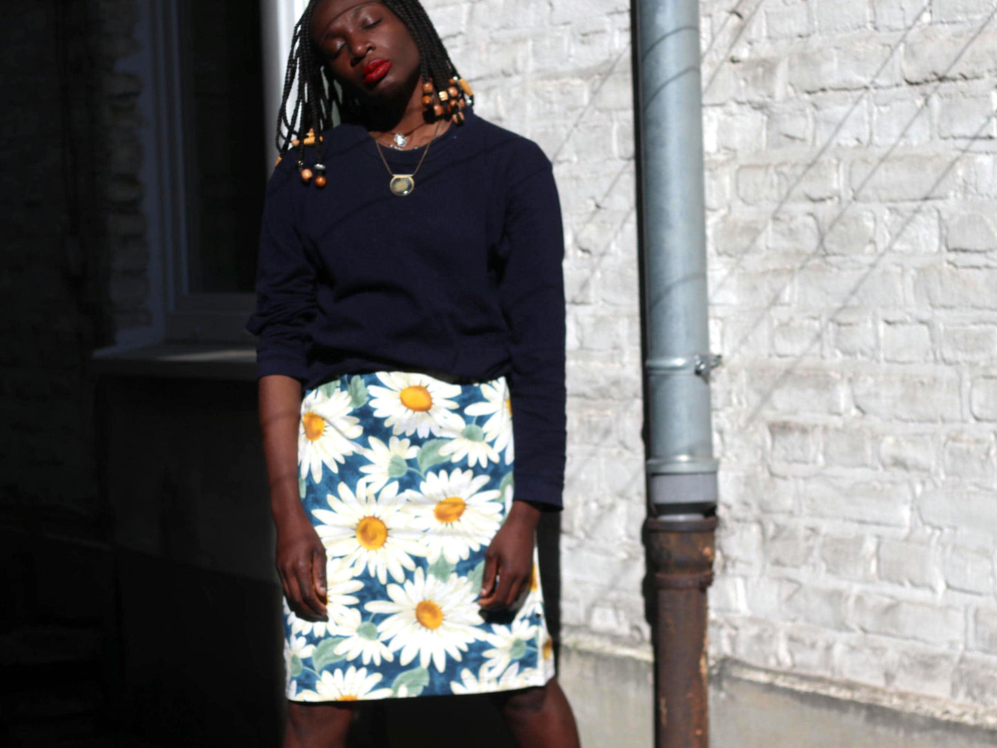 Female model with beaded braids showcasing a 90s vintage daisy print knee-length skirt in white and blue paired with a dark blue sweater is photographed in front of white wall with lined shadows