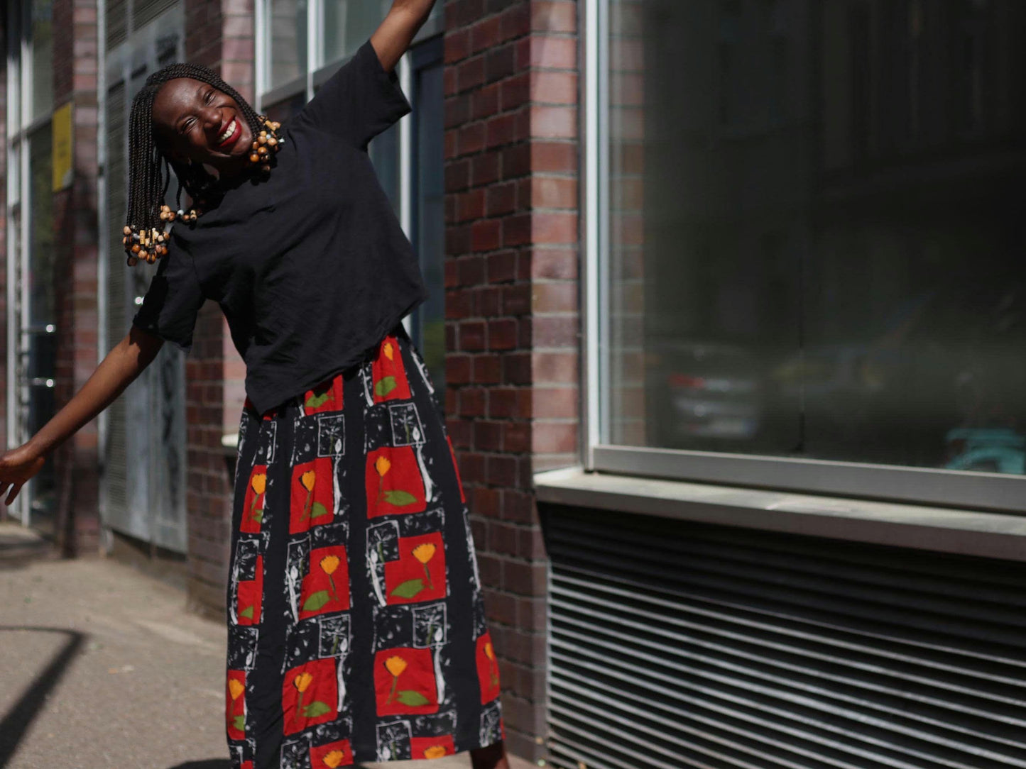 A female model smiles cheerfully in the sunlight wearing a vintage red boho maxi floral skirt, paired with a black top, in front of a brick building and large windows in an urban setting.
