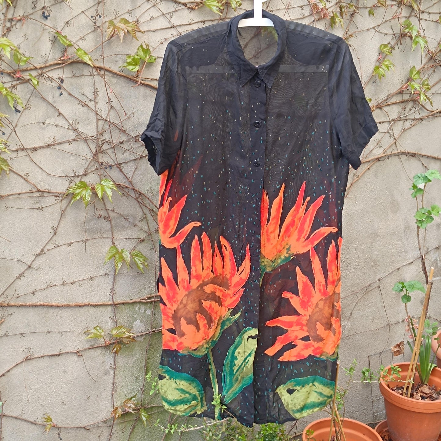 Vintage y2k Black sheer shirt dress with hand-painted orange sunflower design photographed hanging on a clothing line in a garden against a brick wall with browning twines