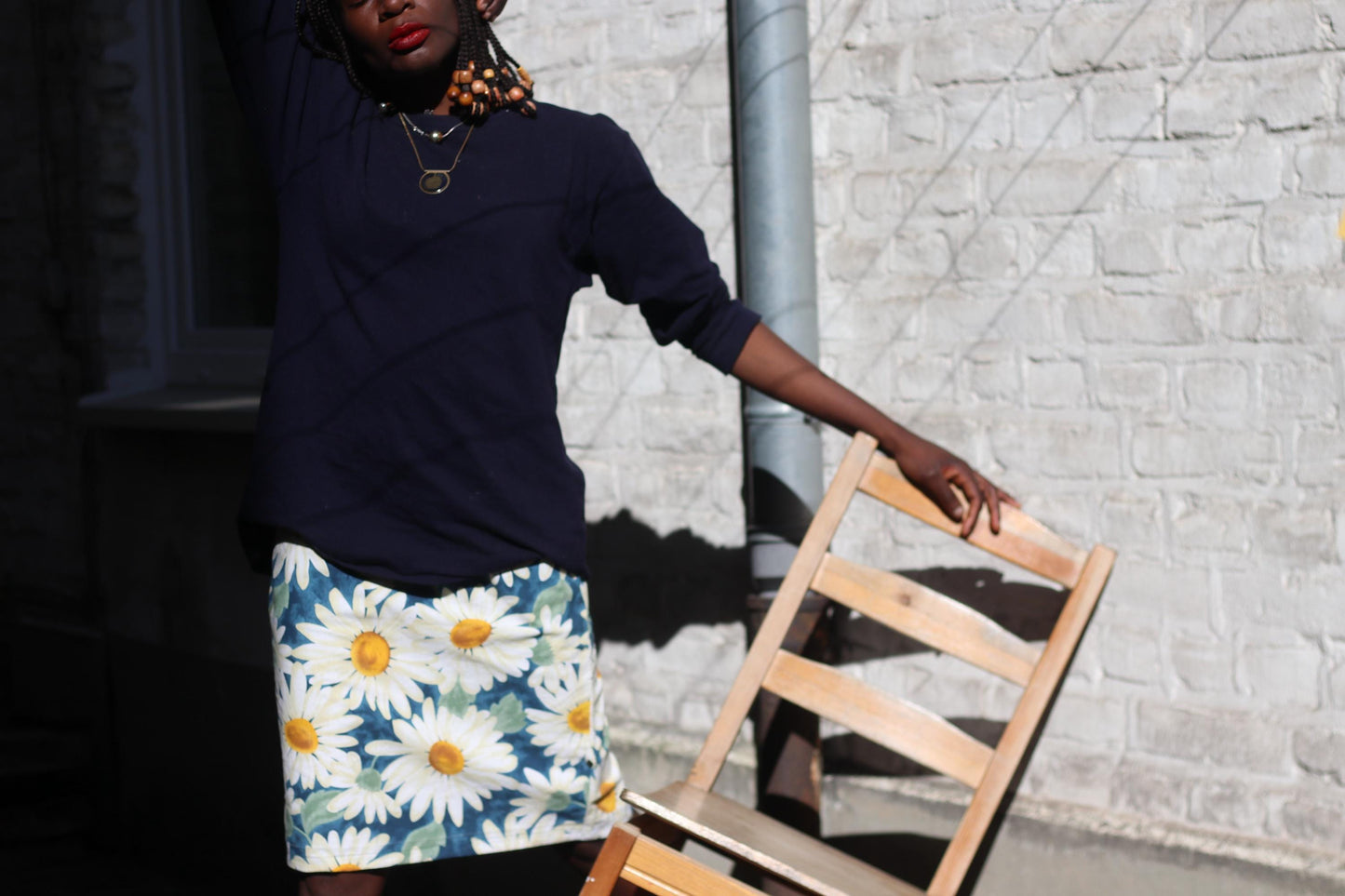 A woman with braided hair and a necklace, wearing a dark top and a vibrant floral skirt, with a brick wall visible in the background and a wooden chair next to her