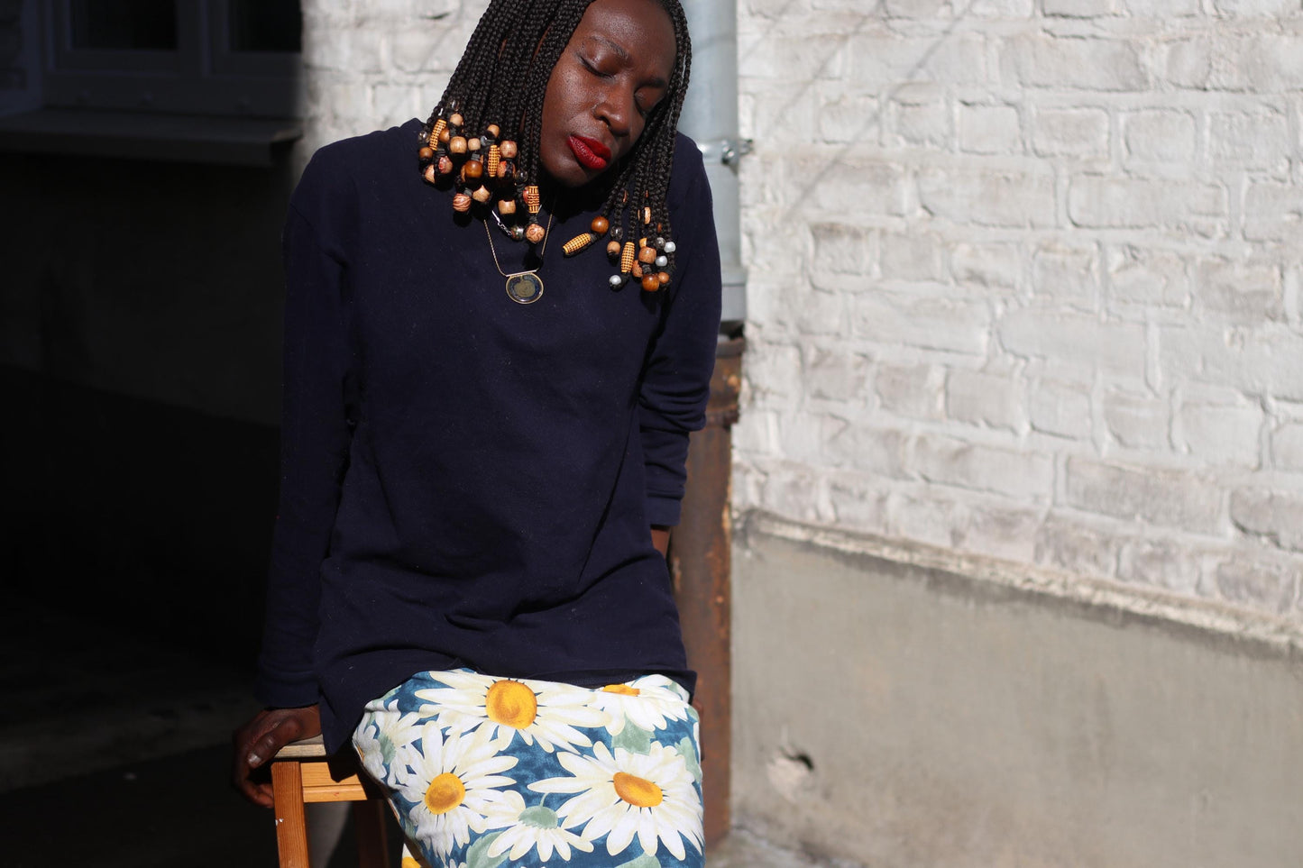 A female model sitting on a chair, showcasing a knee-length skirt with a bold blue and yellow daisy pattern on a white background, paired with a dark long-sleeved shirt.