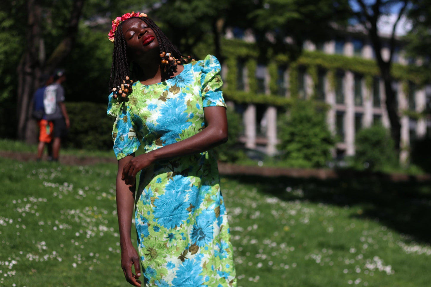 A female model with beaded braided hair wearing a vintage retro dress with bold floral patterns and retro puff sleeves photographed outdoors against a background filled with greenery and daisy flowers