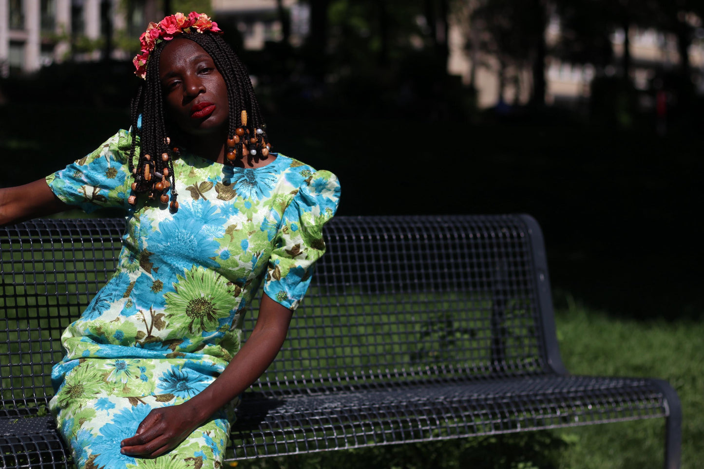 Sitting outdoors on a bench, a person models a handmade-style dress featuring a bold blue and green floral pattern and puff sleeves. They accessorize with beaded braids and a colorful flower crown.