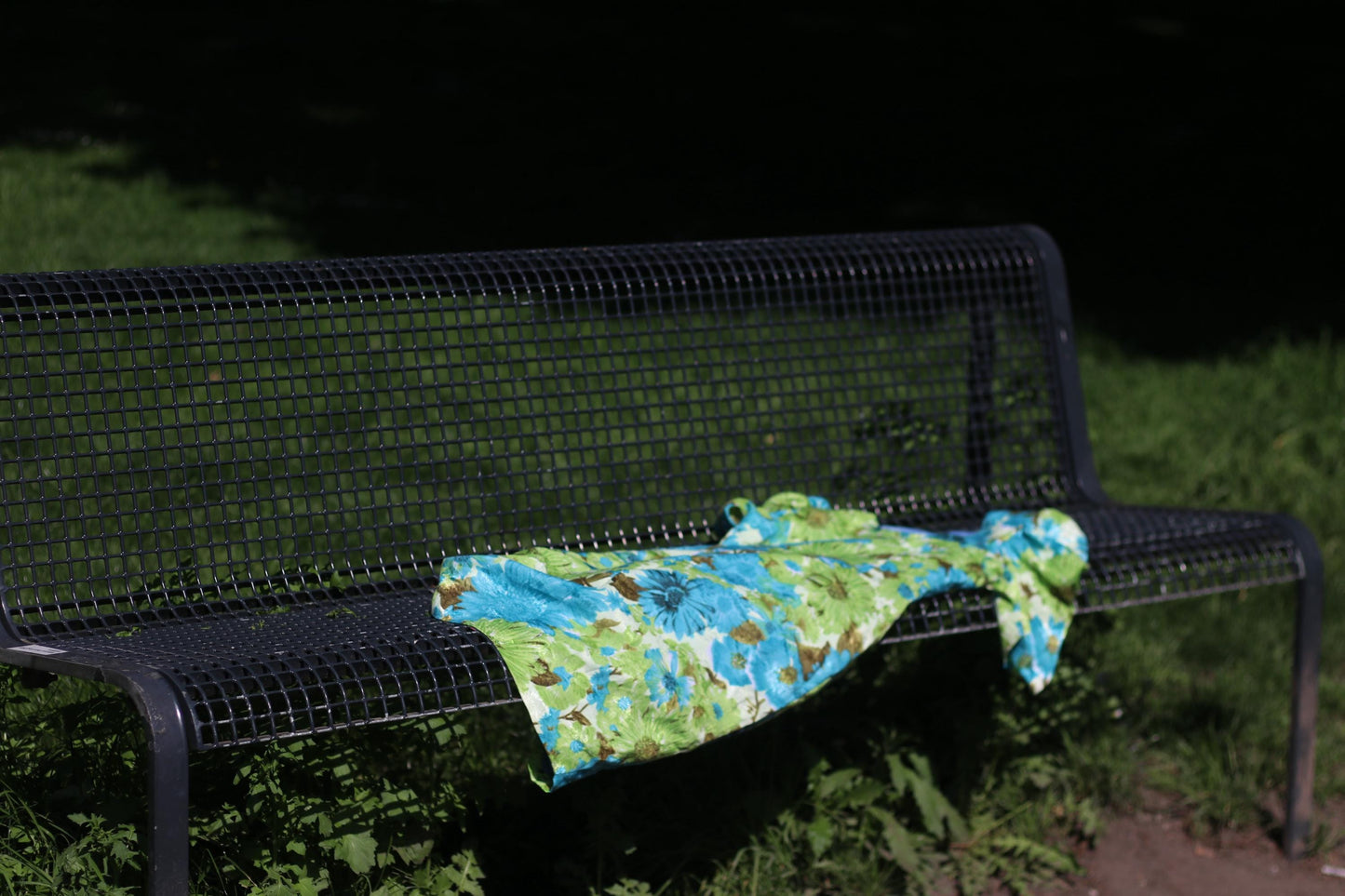 A vibrant vintage 70s dress with a blue and green floral pattern rests on a park bench. The dress features short, puffed sleeves and a flowing design, showcasing its unique handmade style.
