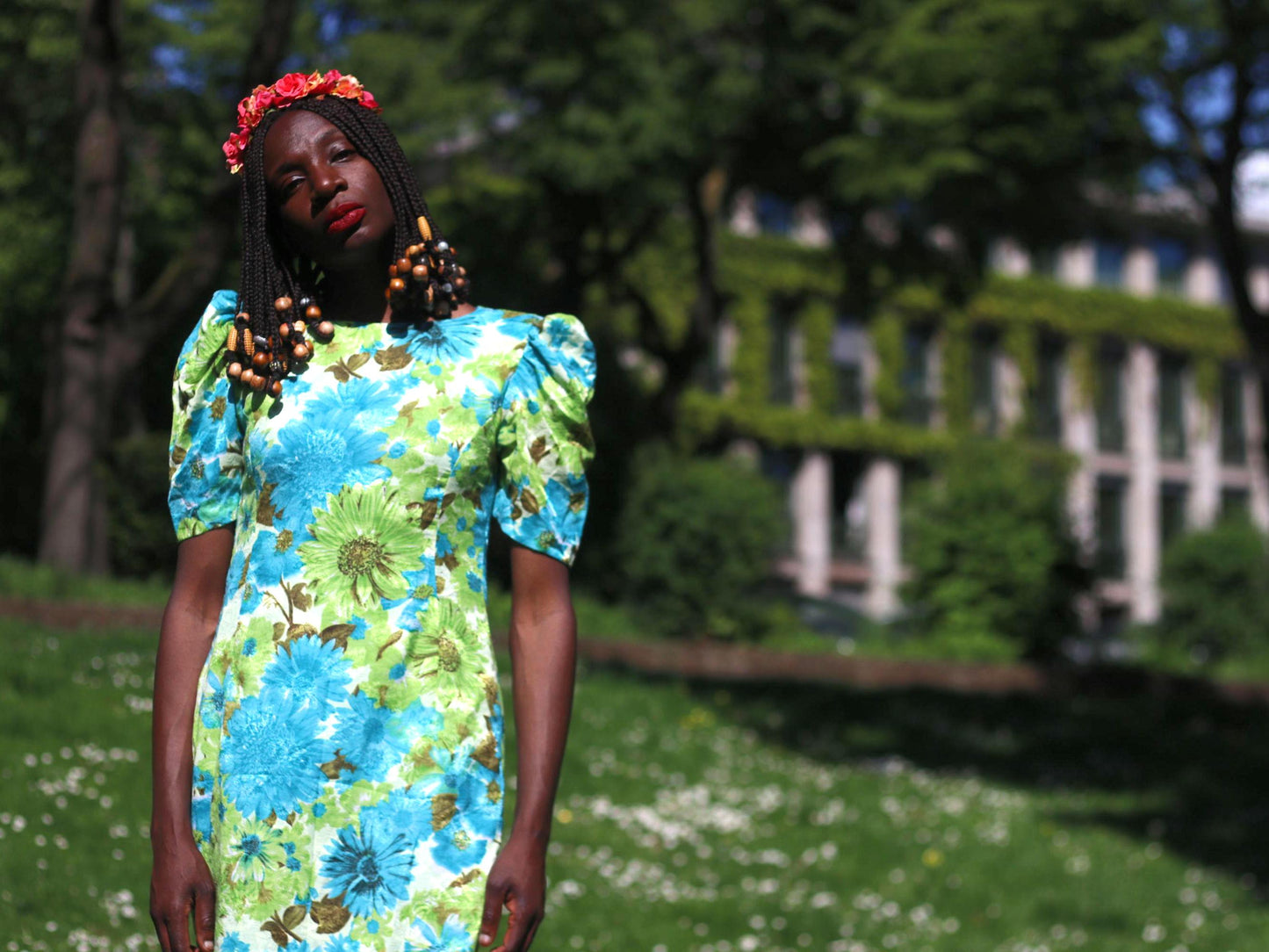 A person with dark skin and long beaded braids, wearing a vibrant blue and green floral dress with short, puffed sleeves and a colorful floral headpiece, is photographed outdoors against a background with greenery and historic looking buildings