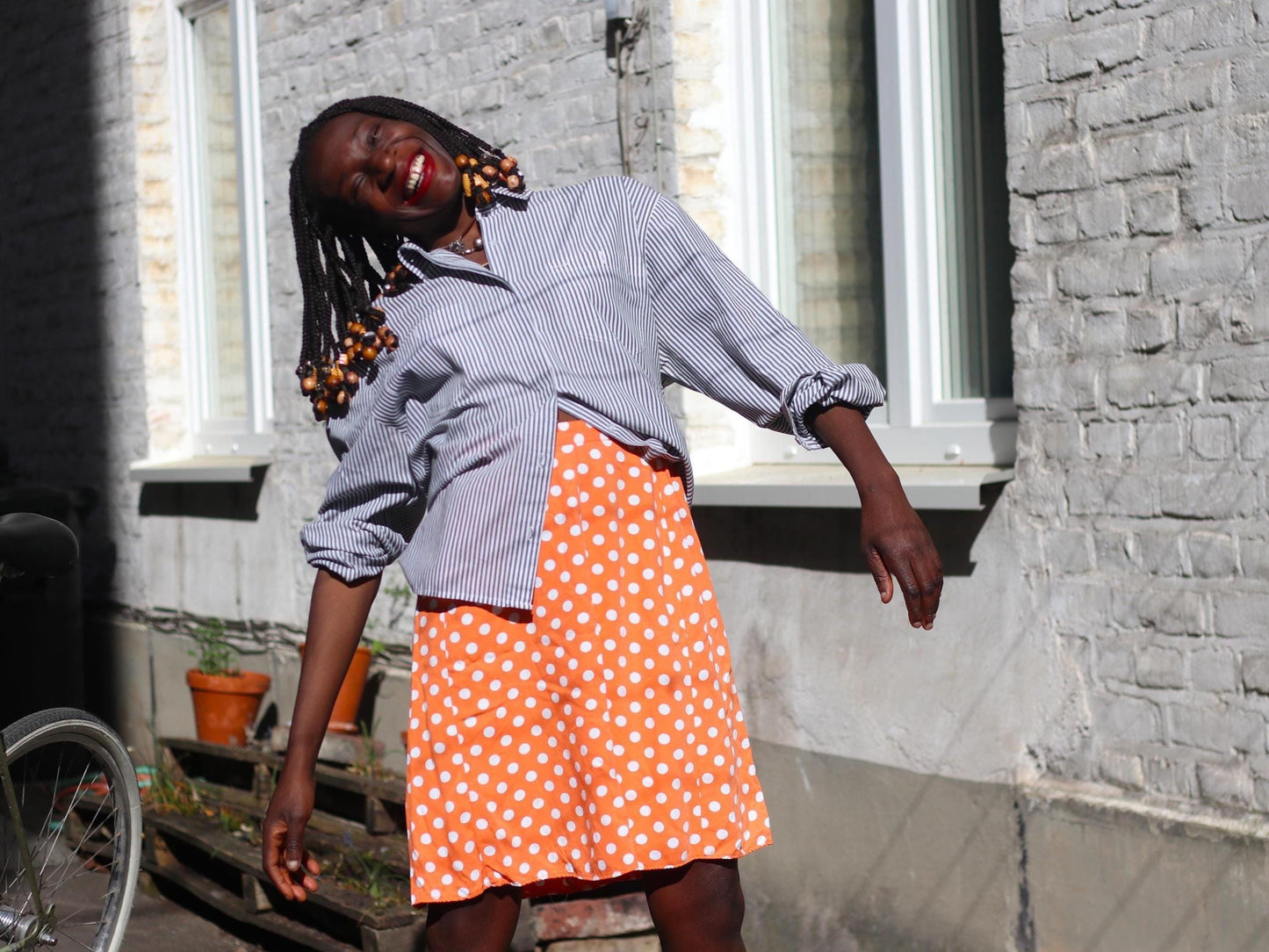 Model of colour in a vintage Parisian chic orange skirt with Polka Dotted paired with blue striped blpuse. The image is shot outdoors and she stands Infront of a sunlit bricked wall, the vibe is summer fun a la Amelie Poulain