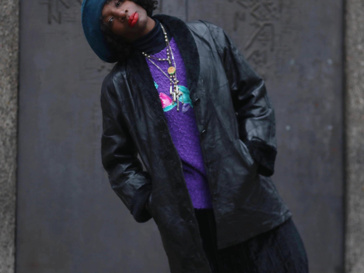 90s Black patchwork leather coat by Anne De Lancay with visible seams, layered over a purple sweater and dark skirt, worn by a female model with layered necklaces and a dark hat. Gray textured wall in the background
