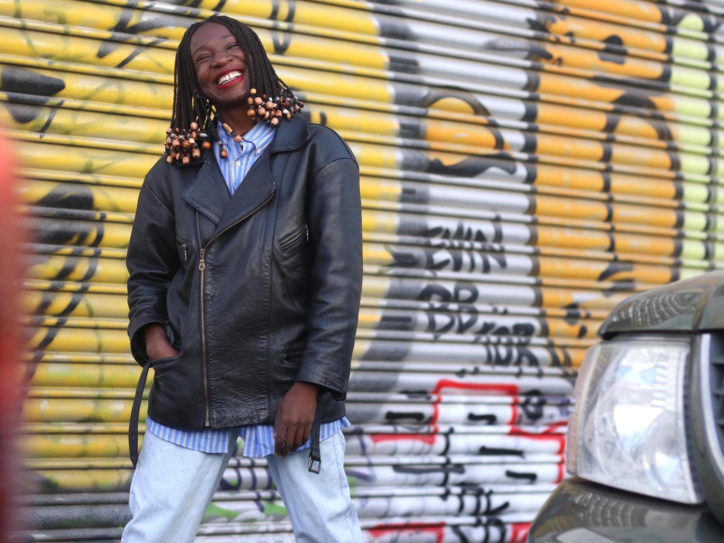 Female model with braids wearing a black vintage Rock and roll style leather jacket from the 90s is photographed outdoors against a graffiti covered wall with smiling. She has a striped blue shirt underneath paired with light blue jeans.