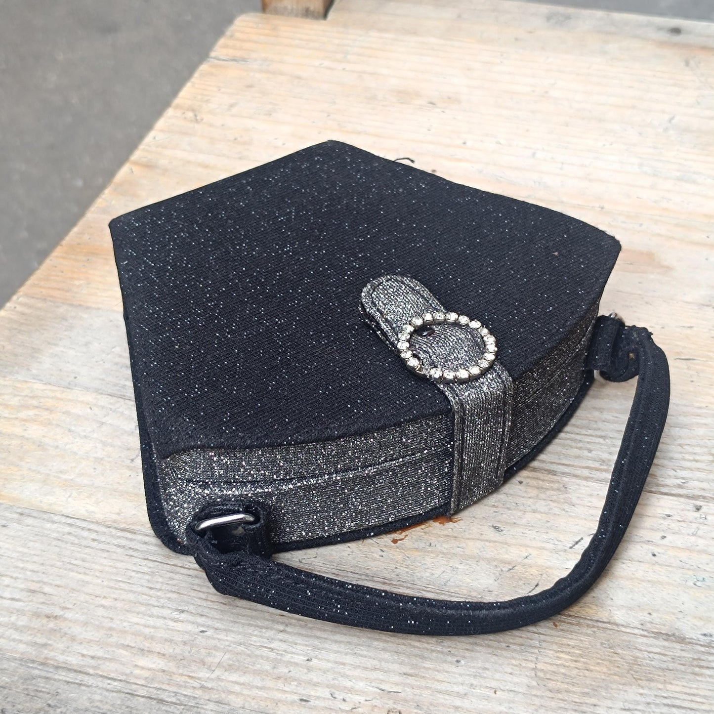 Close-up of a black glitter y2k mini bag with a circular faux-bling embellishment on the tab closure. The compact vintage bag's top handle and pentagon shape are also visible against a rustic wooden backdrop."