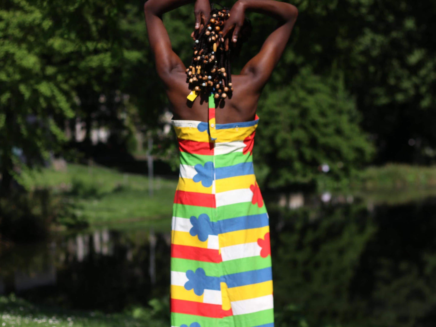 Back view of a woman wearing a bold 1970s floral halterneck dress with primary-colored stripes in a sunny park