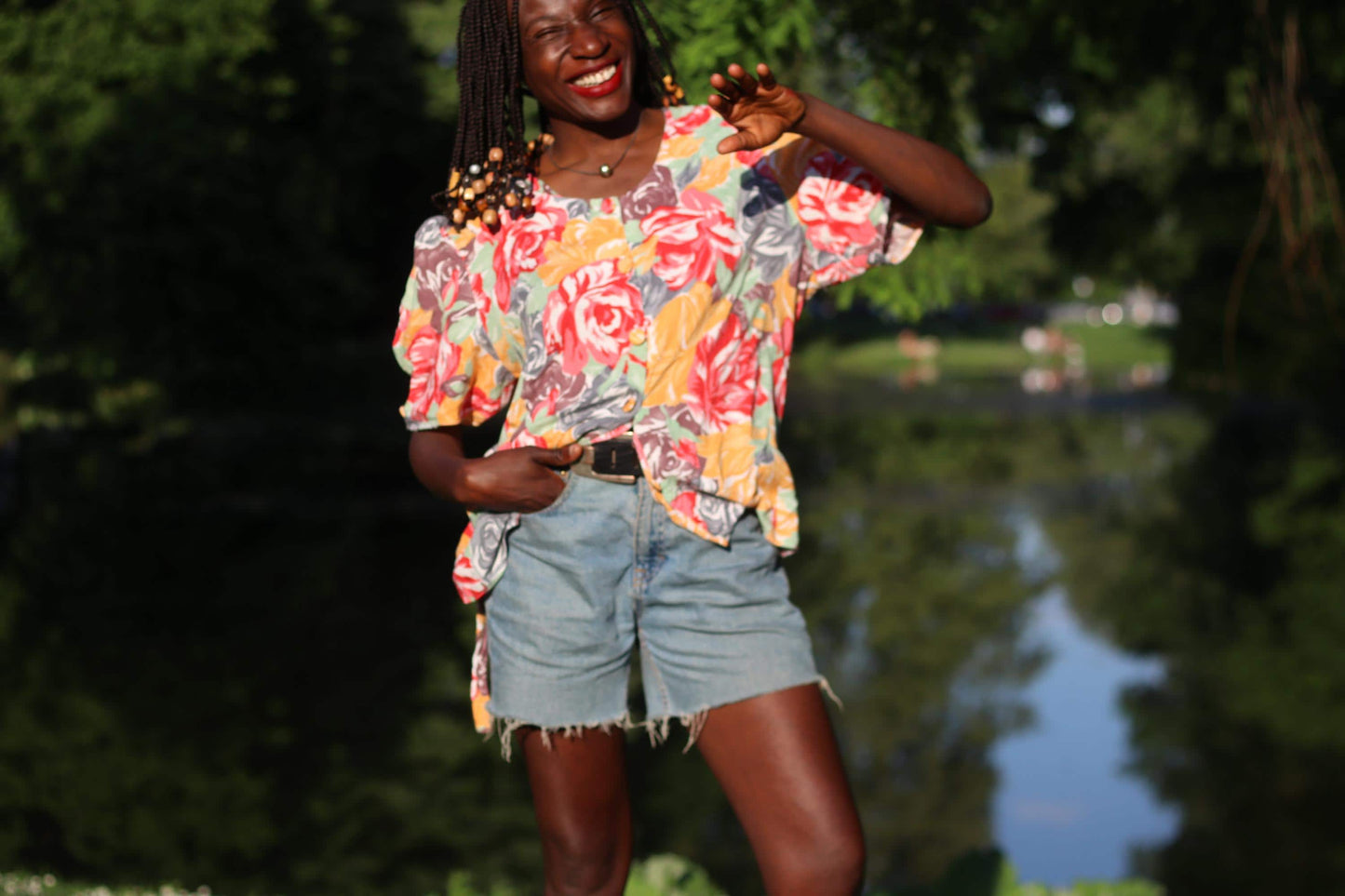 Smling model with beaded braids wearing a vintage floral blouse with puff sleeves combined with blue denim shorts is photographed sitting in the grass, looking into the camera on a sunny day. Background is greenery