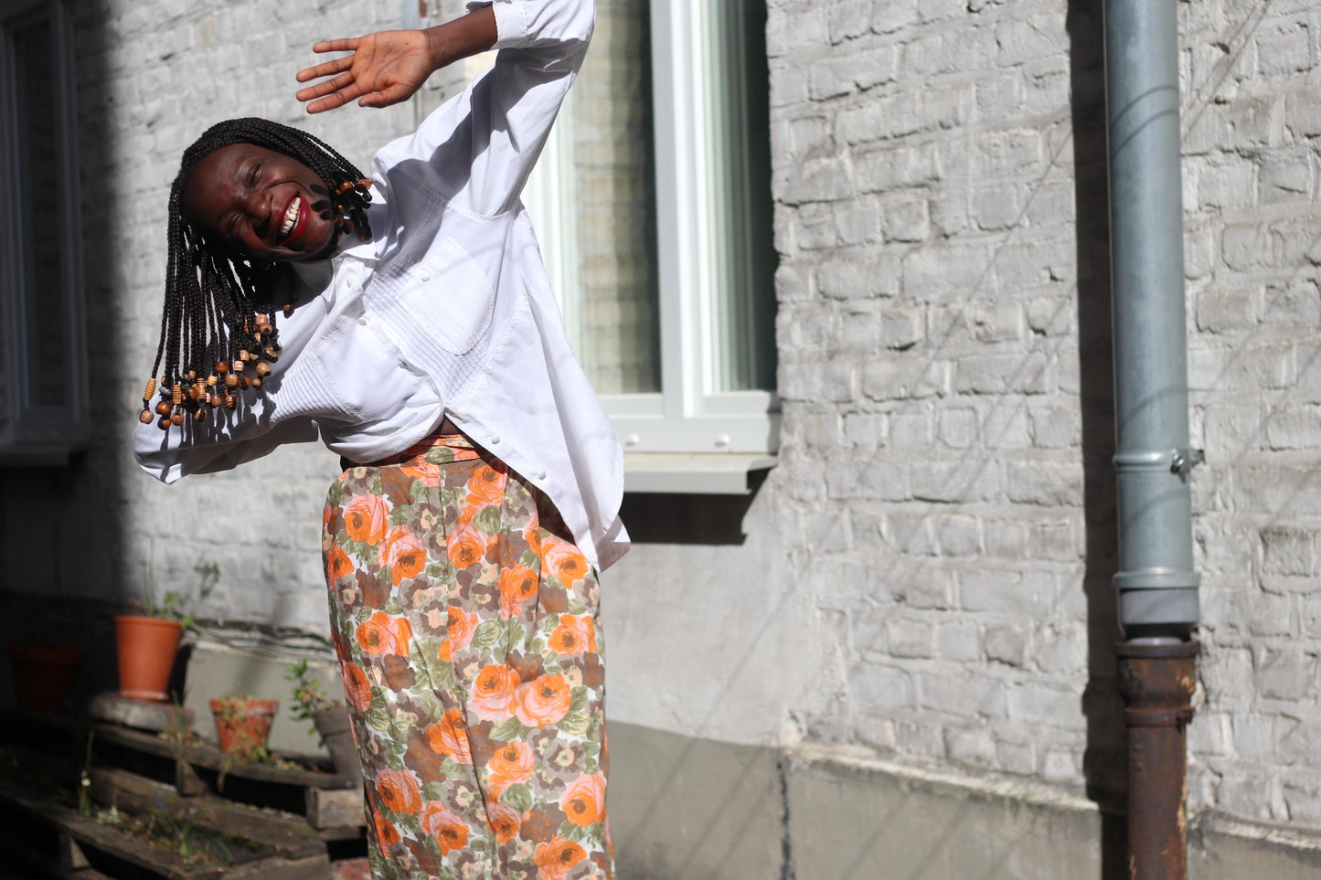 Smiling Female model wearing an 80s Floral Skirt in orange and brown styled with a pinstriped button-up in photographed outdoors against a sunlight white bricked wall. Her hands are up as if to wave