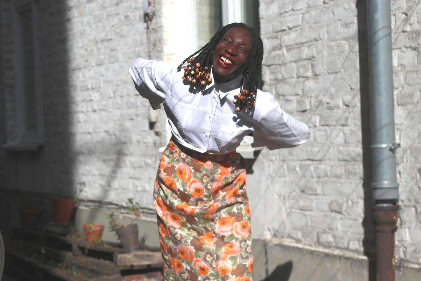 Smiling female model with beaded braids wearing an orange floral skirt from the 80s photographed outdoors in a garden Infront of a sunlit bricked white wall. The cheerful image focus on the maximalist print on the vintage skirt, evoking the joy of summer.