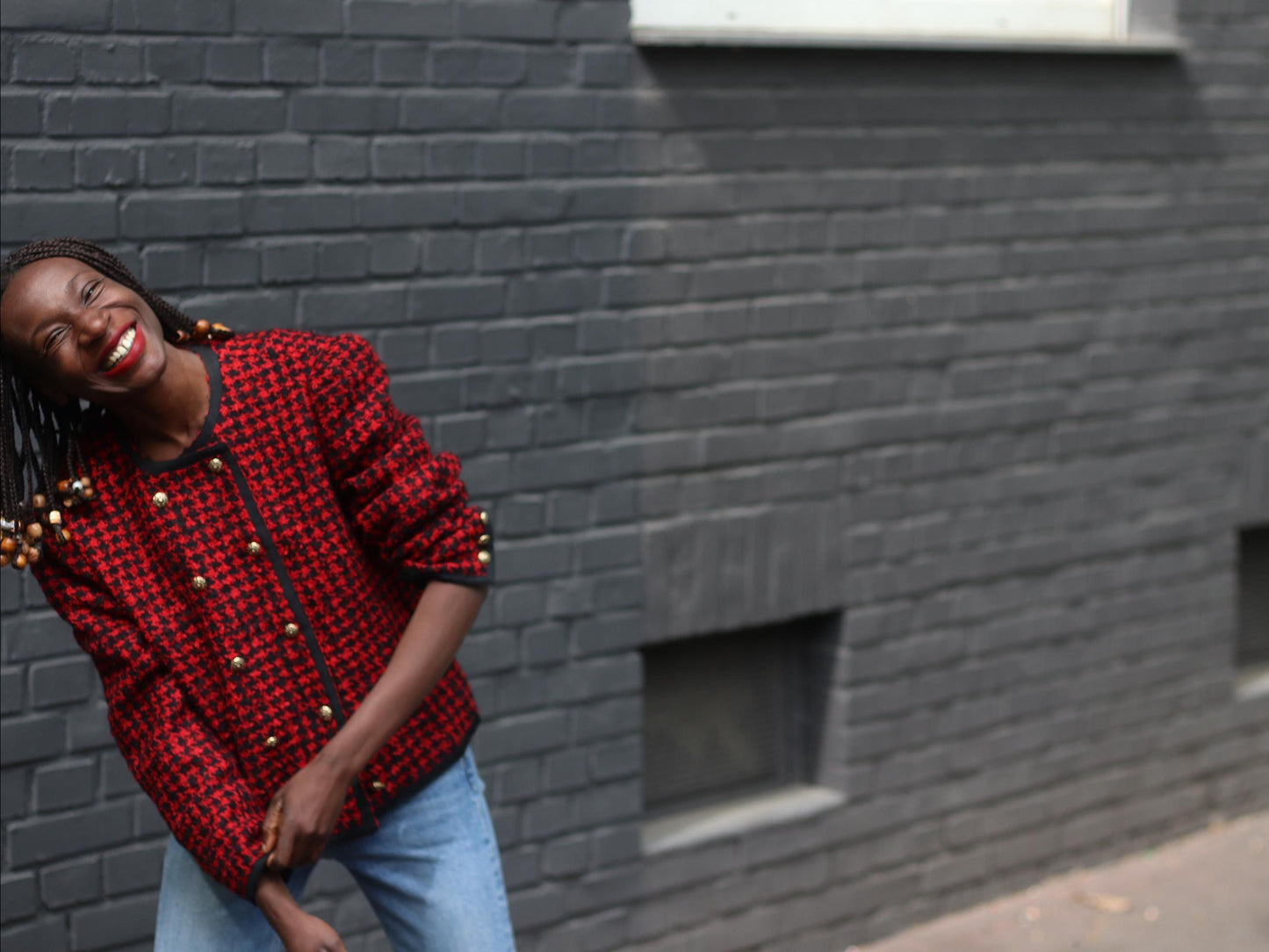 Vintage red houndstooth Basler wool blazer with gold buttons showcased on a smiling model of African heritage with beaded braids, full front view against grey bricked walled background.